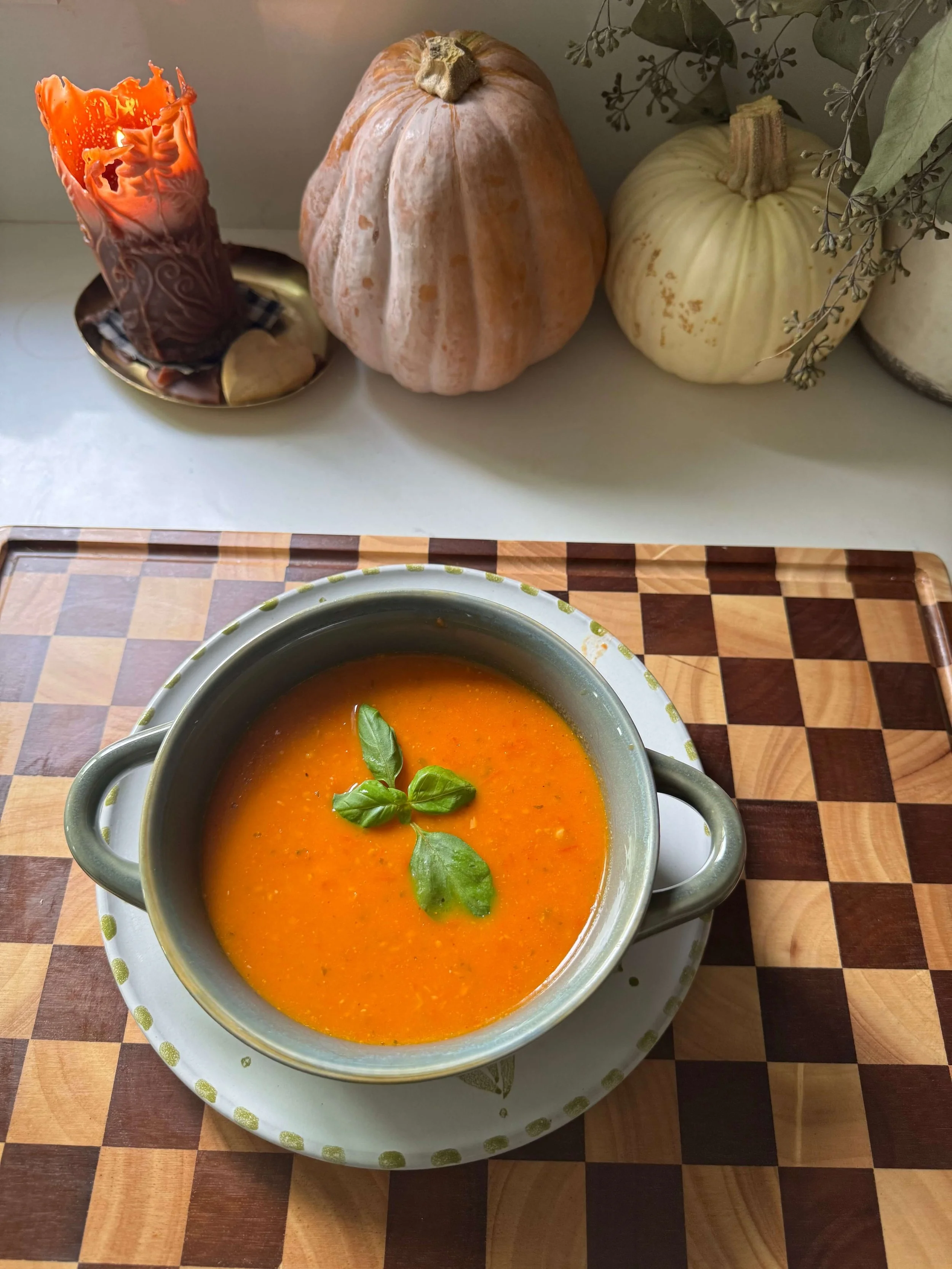 tomato soup on counter on checkered cutting board pumpkins on kitchen countertop with basil on soup in blue bowl