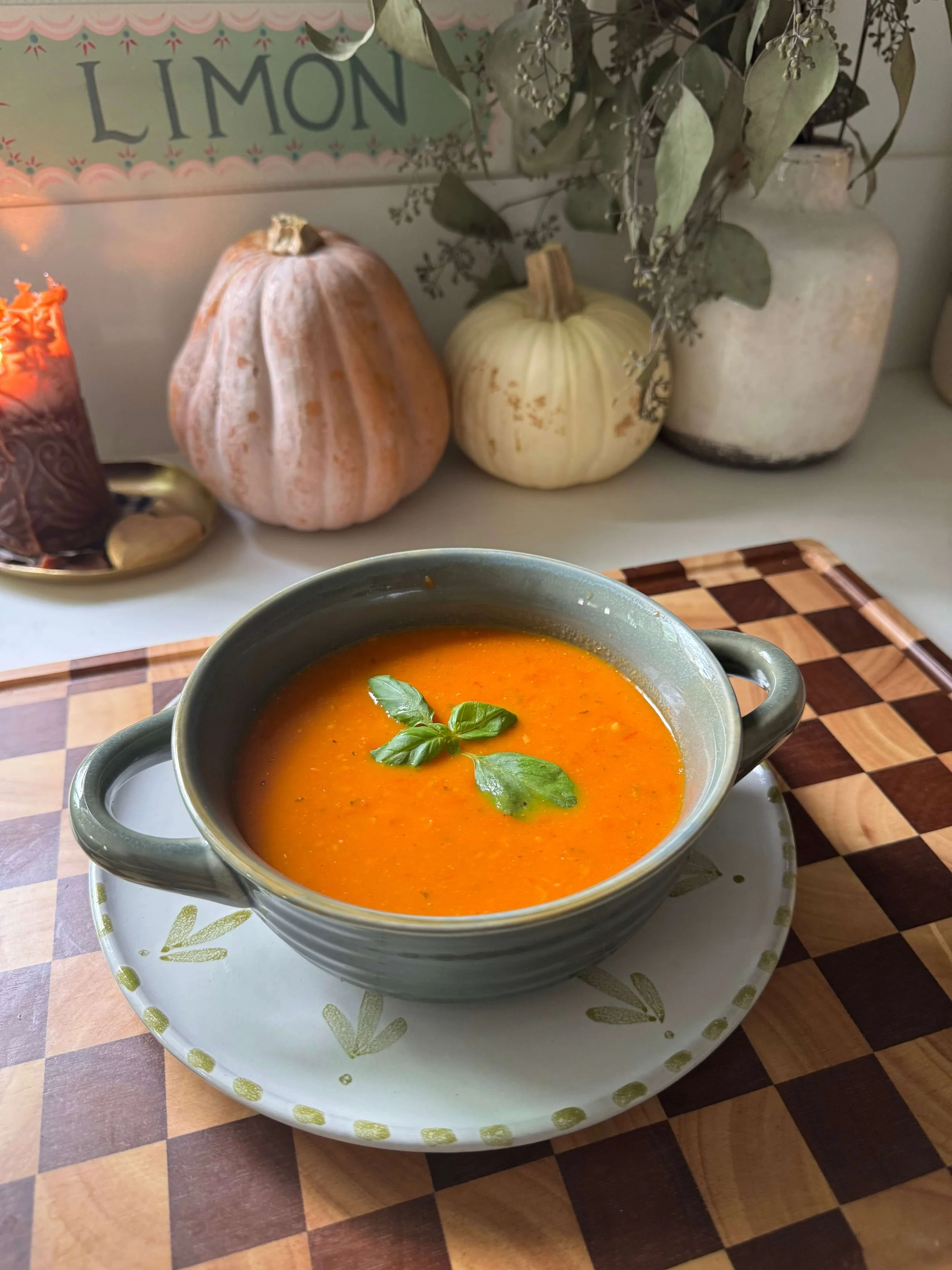 tomato soup on counter on checkered cutting board pumpkins on kitchen countertop with basil on soup in blue bowl