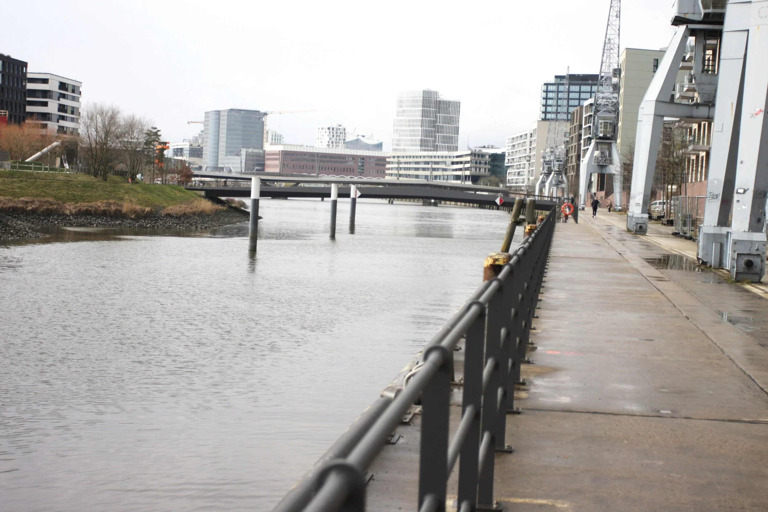 Stadtansicht mit Fluss, Brücke und modernen Gebäuden, dazu eine Promenade mit Geländer und Fußgängern.
