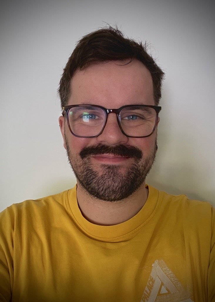A man with short brown hair, glasses, a full beard, and earrings, smiling at the camera against a plain white wall.