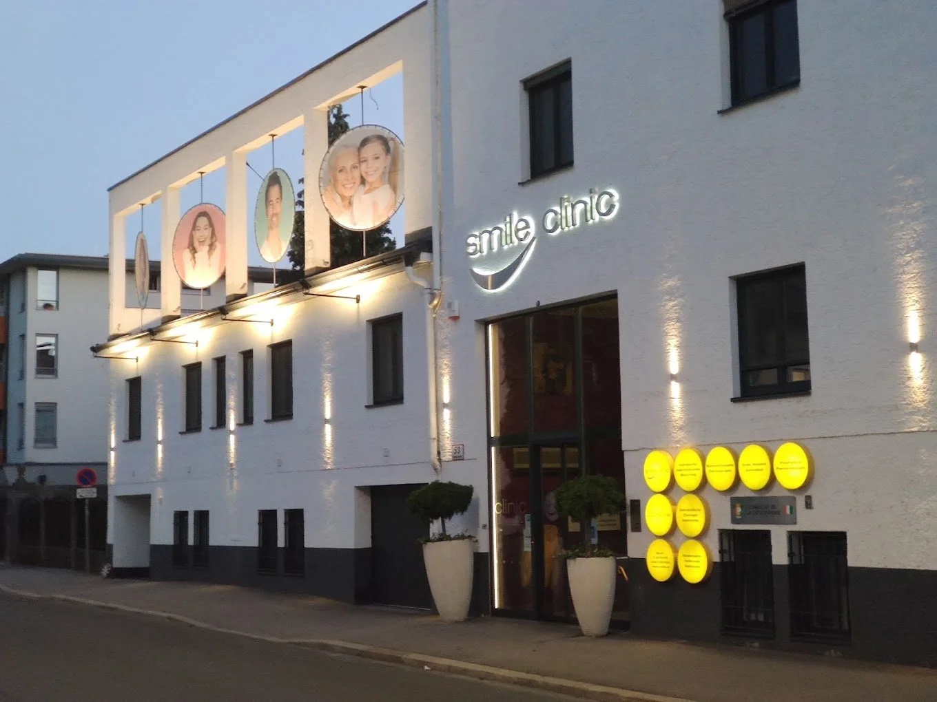 Das Bild zeigt das Äußere eines modernen Zahnärztezentrums namens 'smile clinic' an einem Abend, mit beleuchteten Fotos von lachenden Menschen an der Fassade, gelben Hinweisschildern und großen Pflanzkübeln vor dem Eingang.