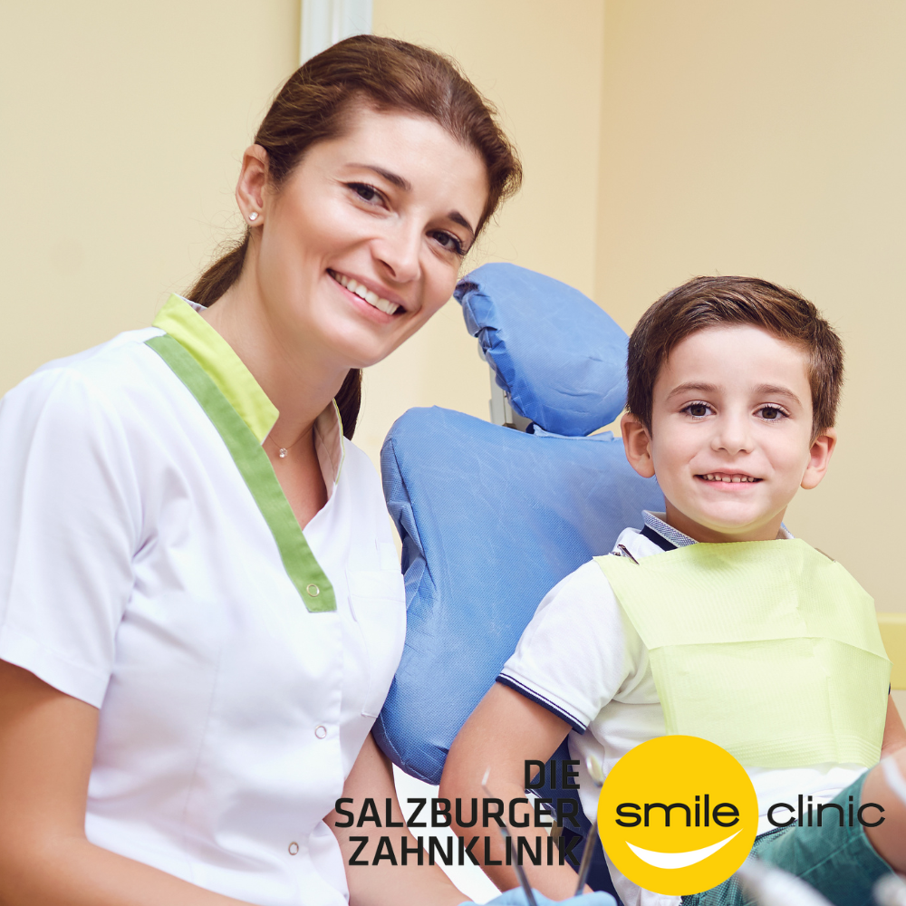 Ein strahlendes Mädchen und ein Junge sitzen in einem Behandlungsraum der Salzburger Zahnarztklinik, beide tragen Schutzkittel, das Mädchen trägt Ohrringe und hat braune Haare, der Junge hat braune Haare und trägt einen weißen und blauen Pullover.