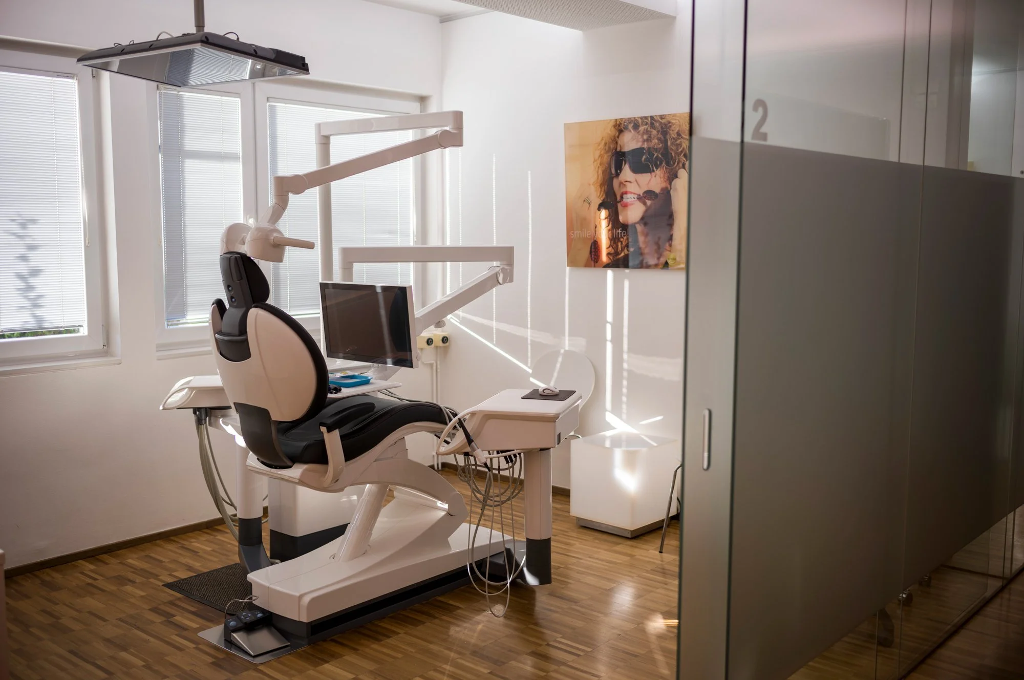Säuglingsstuhl in einem Behandlungsraum einer Zahnarztpraxis, mit medizinischen Geräten und Monitoren, neben einer Wand mit einem Porträt einer Frau mit Sonnenbrille.