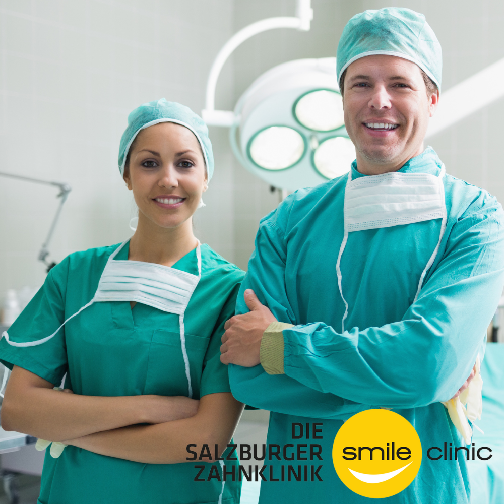 Zwei Zahnärzte in Operationskleidung stehen in einer Klinik, lächeln und blicken in die Kamera, im Hintergrund Operationslampen.