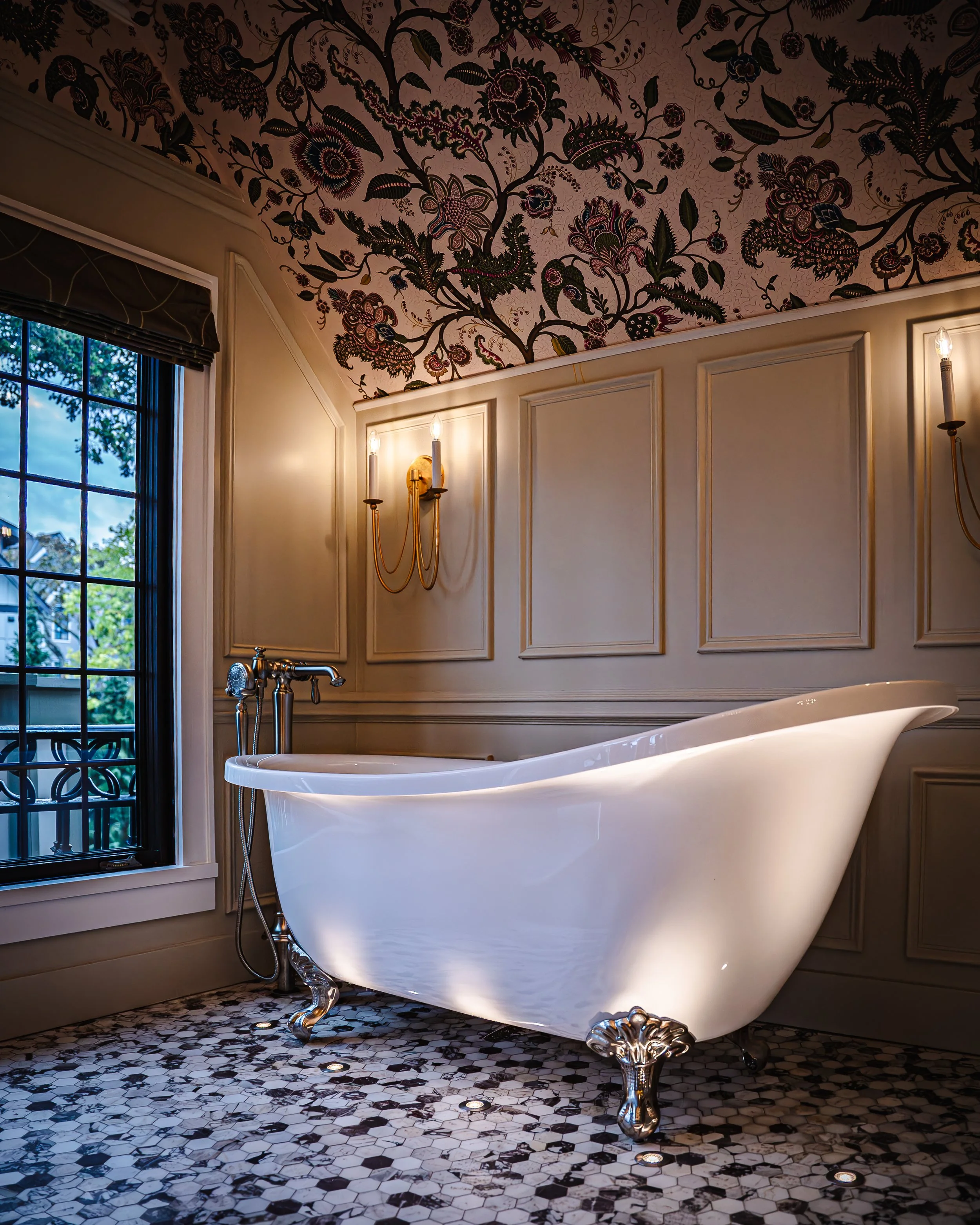 Brass wall sconces and recessed floor lights in a renovated bathroom with a clawfoot tub and floral wallpaper
