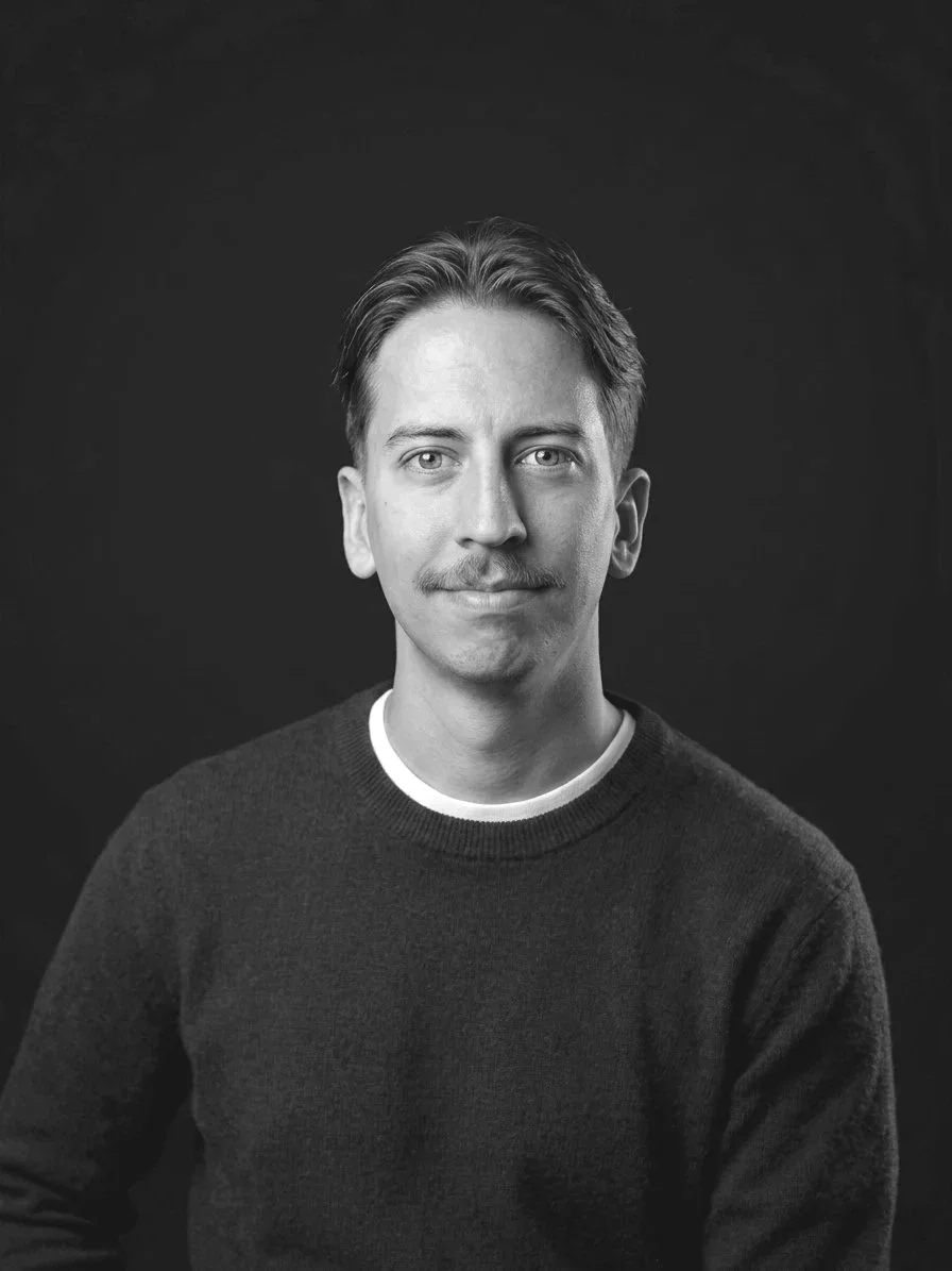 Black and white portrait of a man with light skin, short hair, mustache, wearing a dark sweater, sitting against a dark background.