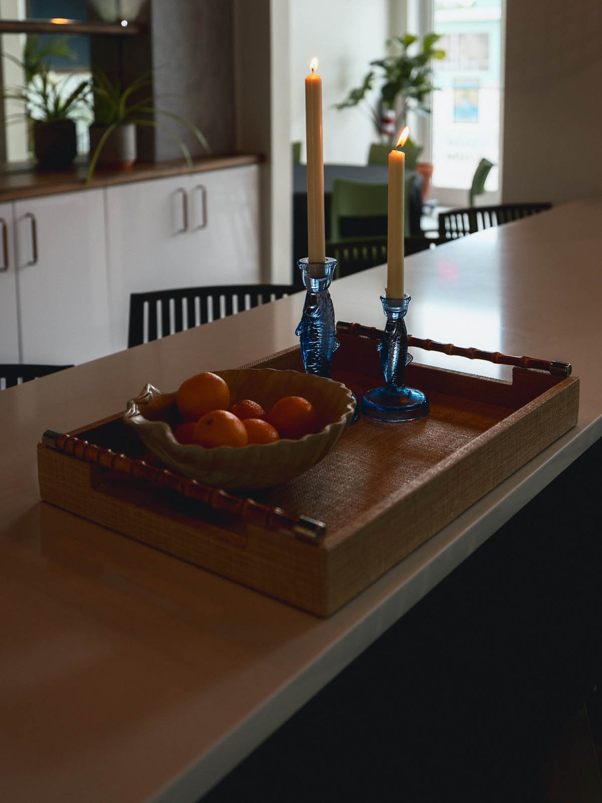 A wooden tray on a table holding a ceramic bowl of oranges and a blue glass candle holder with two lit candles in a cozy, well-lit home interior.