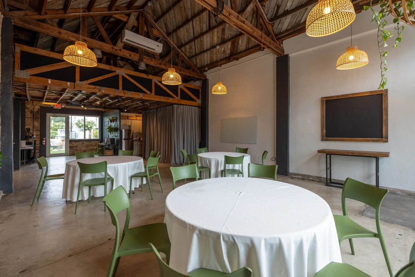 Interior of a modern event space or restaurant with round tables covered in white tablecloths, green chairs, exposed wooden beams, pendant lighting, and a chalkboard on the wall.