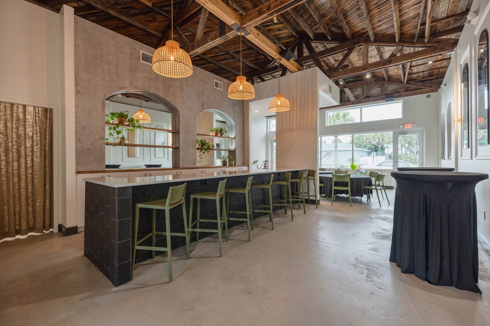 Interior of a modern restaurant or cafe with a high wooden ceiling, hanging wicker lights, a bar with green chairs, round tables with black tablecloths, and large glass windows letting in natural light.