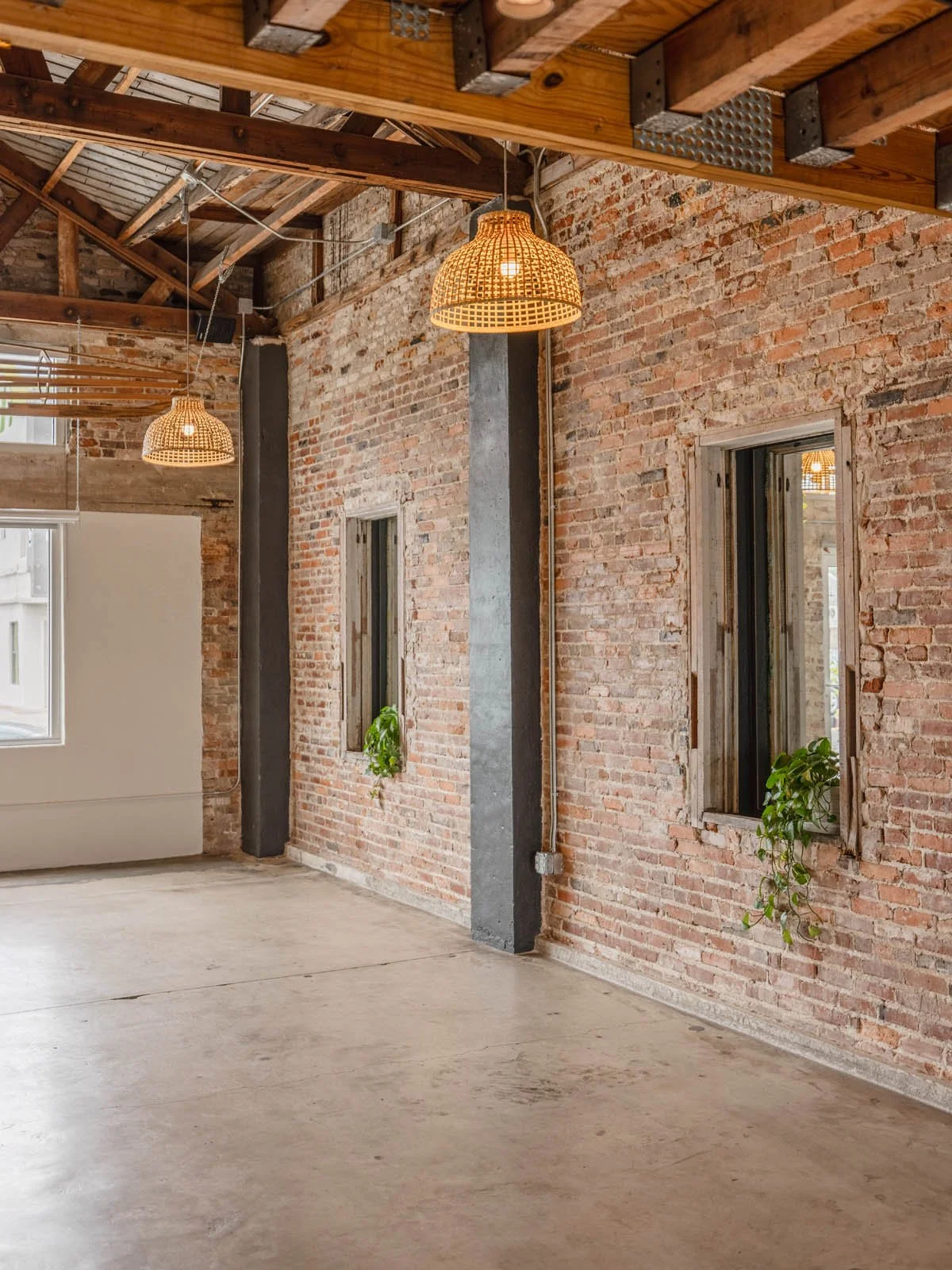 Interior view of a space with exposed brick walls, small windows with plants, wooden ceiling with hanging rattan light fixtures, and a concrete floor.
