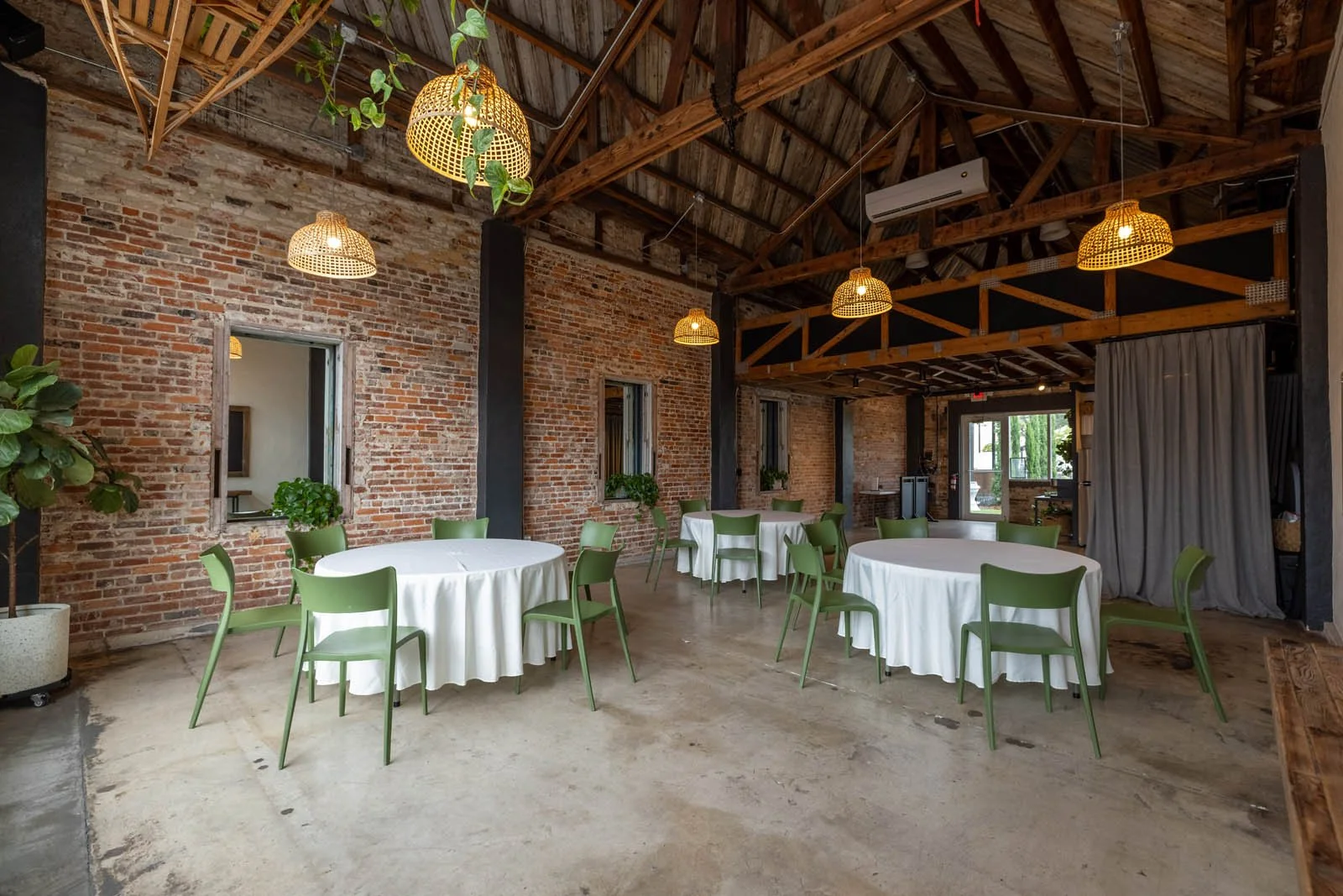 Event space with brick walls, exposed wooden beams, hanging wicker light fixtures, green chairs, and round tables with white tablecloths, decorated for a gathering.