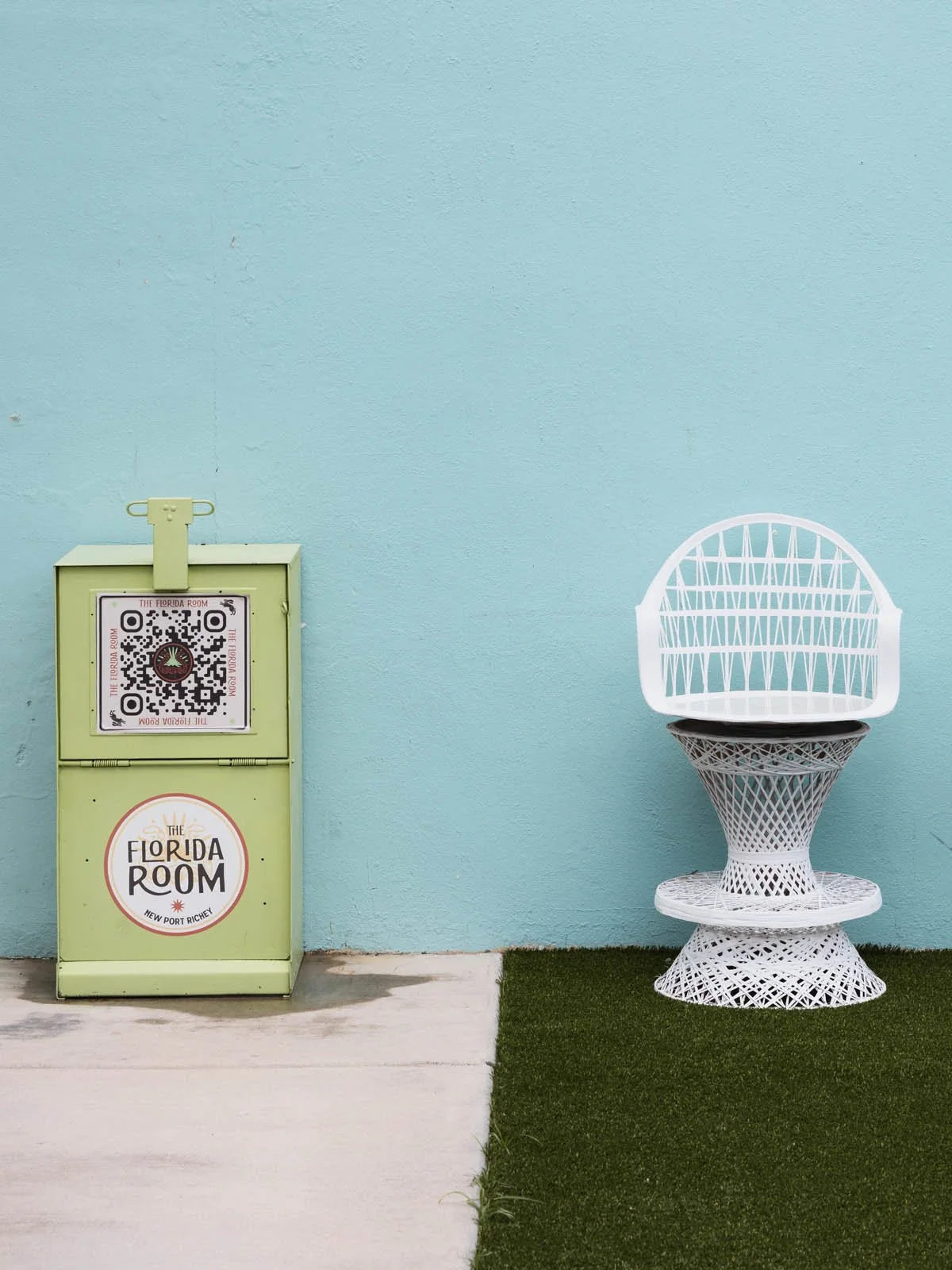 A light green stand with a QR code and sign reading 'The Florida Room, New Port Richey' next to a white decorative chair with a lattice design on a turf and concrete surface against a light blue wall.