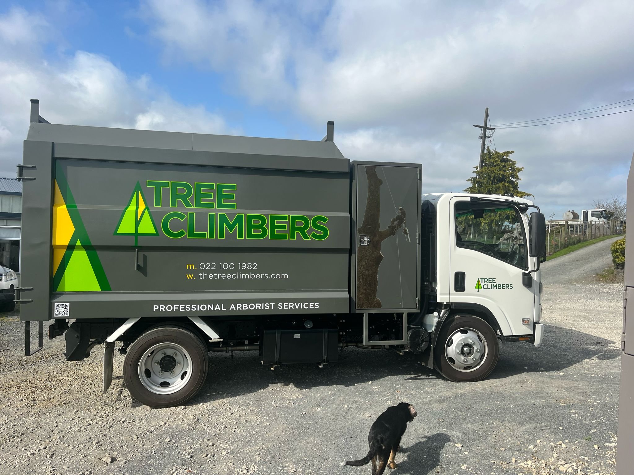 A truck for Tree Climbers company parked outdoors with a black dog nearby, featuring company branding and contact information on the side.