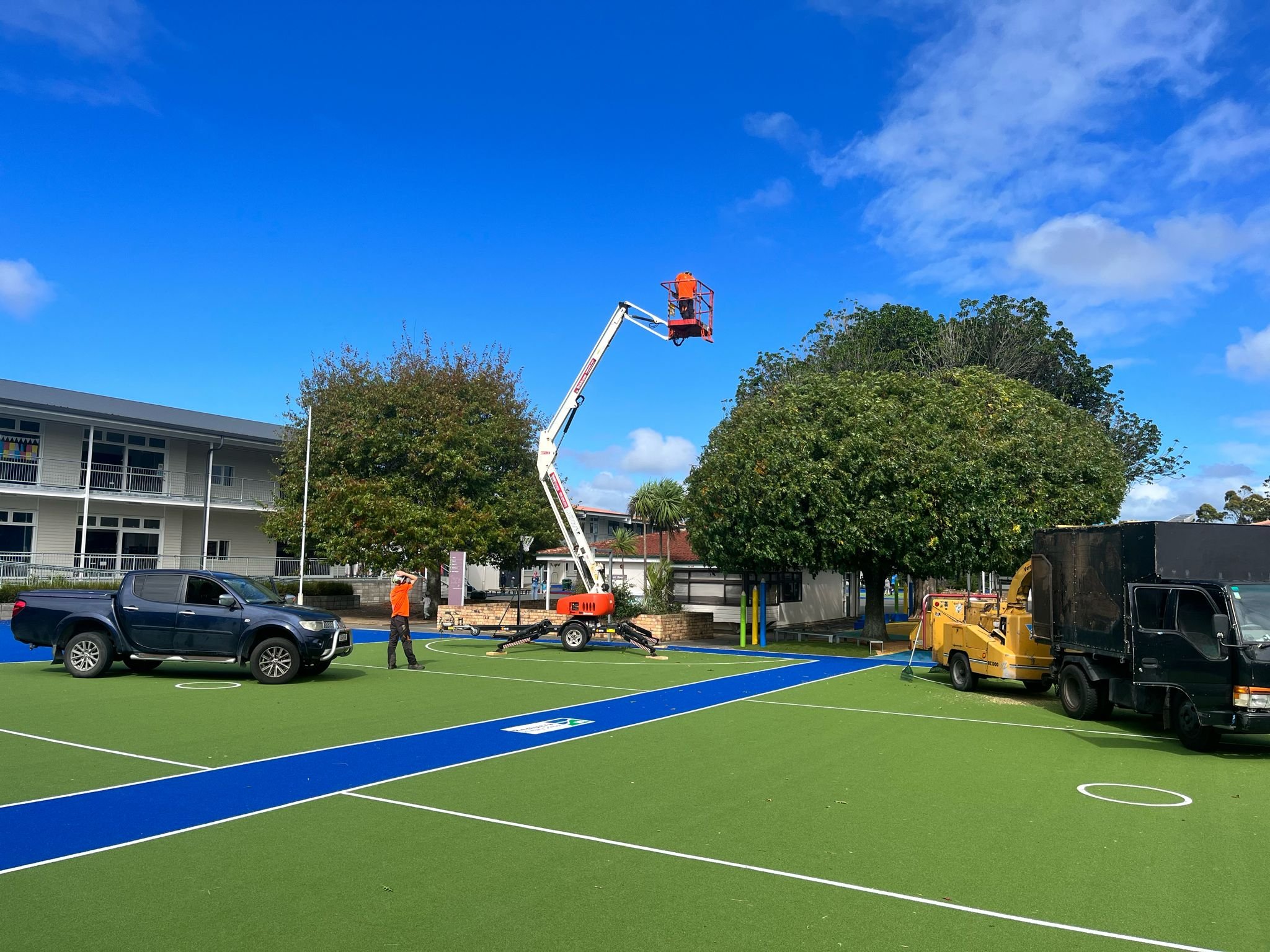 Workers installing a basketball hoop on an outdoor court with a blue and green synthetic surface, surrounded by trees, trucks, and a building under a partly cloudy sky.