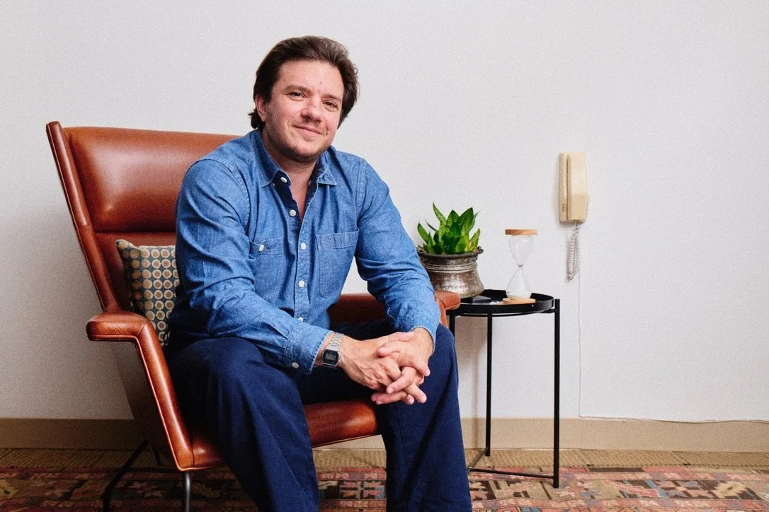 Man sitting in a brown leather chair in a room with white walls, next to a small black side table with a plant, an hourglass, and a telephone.