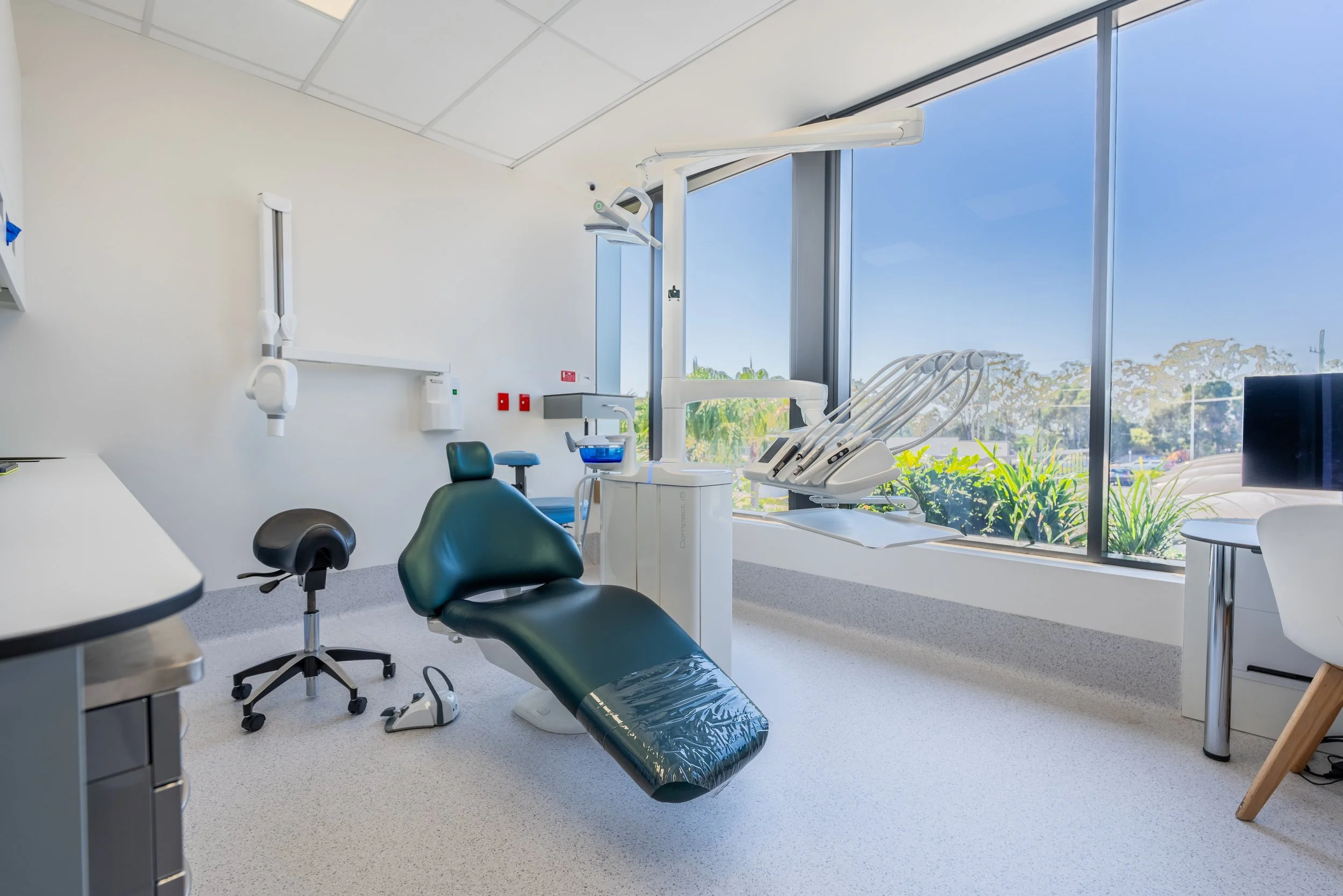 Modern dental clinic treatment room with dental chair, equipment, and large window showing outdoor greenery.
