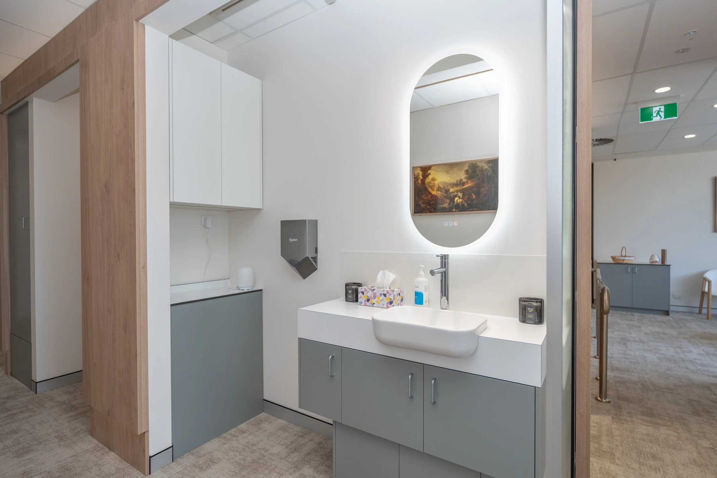 Indoor medical or office facility with sink, mirror, soap dispenser, paper towels, and artwork on the wall.