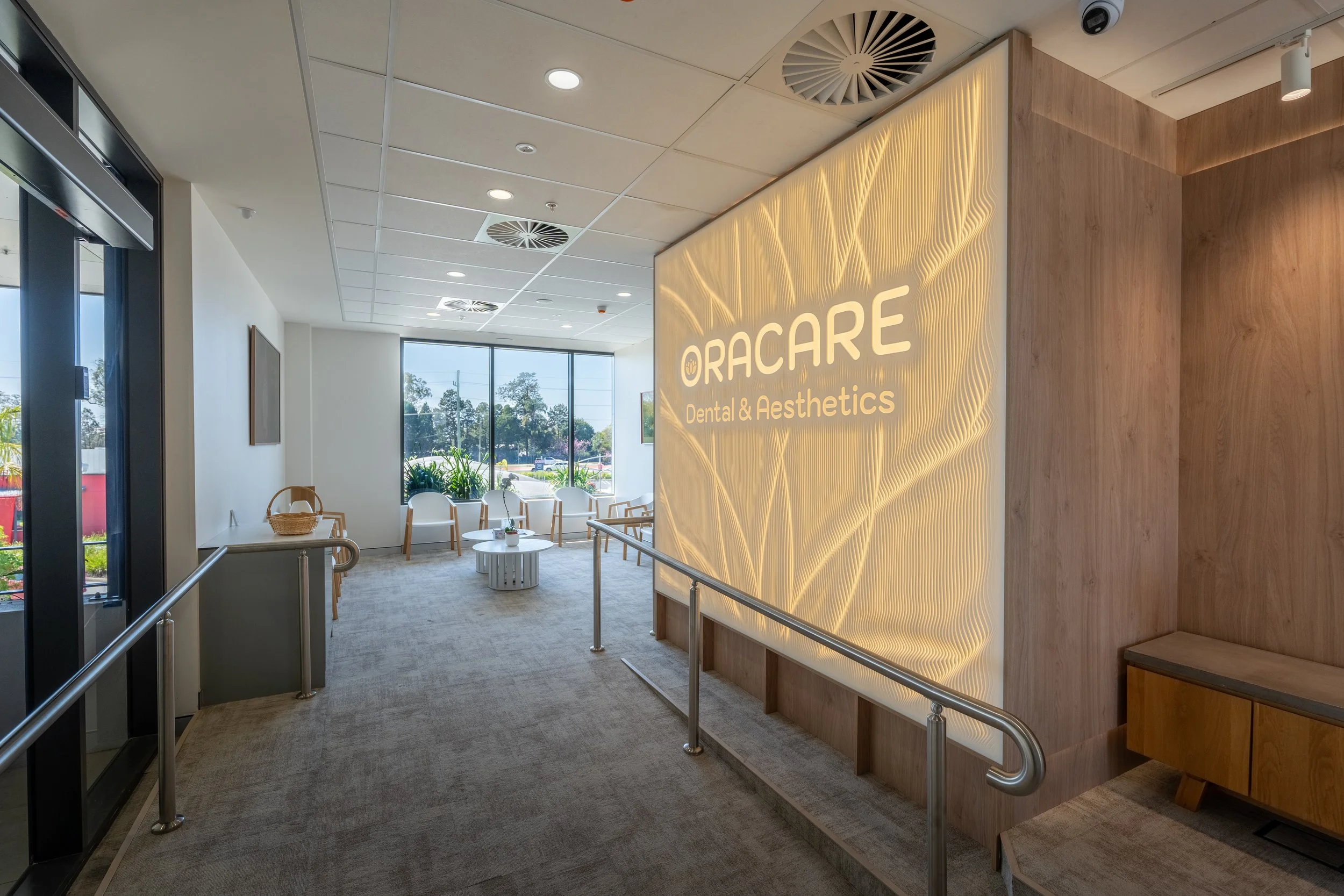 Reception area with sign for Oracare Dental & Aesthetics, seating area with chairs and tables, large window with outdoor view, wood panel wall with illuminated sign.