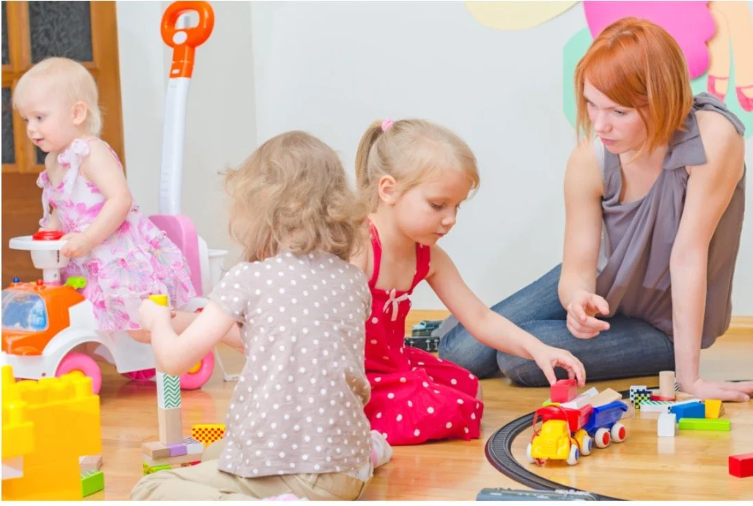 Four young girls, with one adult woman, playing with toys and a train set on a wooden floor in a playroom.