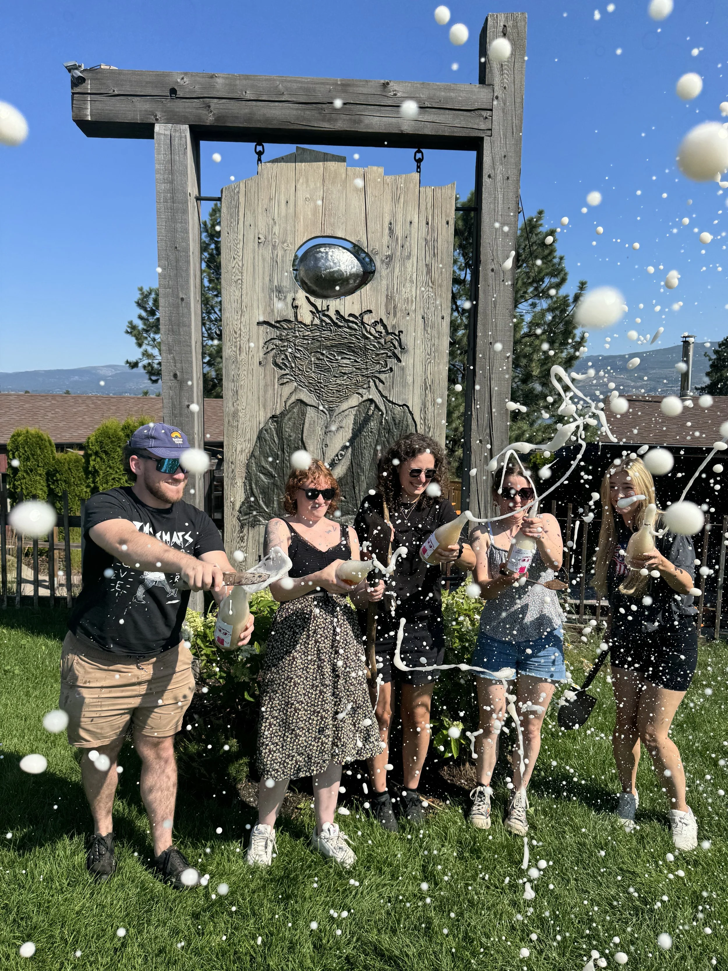 Group of five people celebrating outside with foam bottles, foam flying in the air, and a background of a wooden art piece with a figure and a weather-themed mural under a clear blue sky.