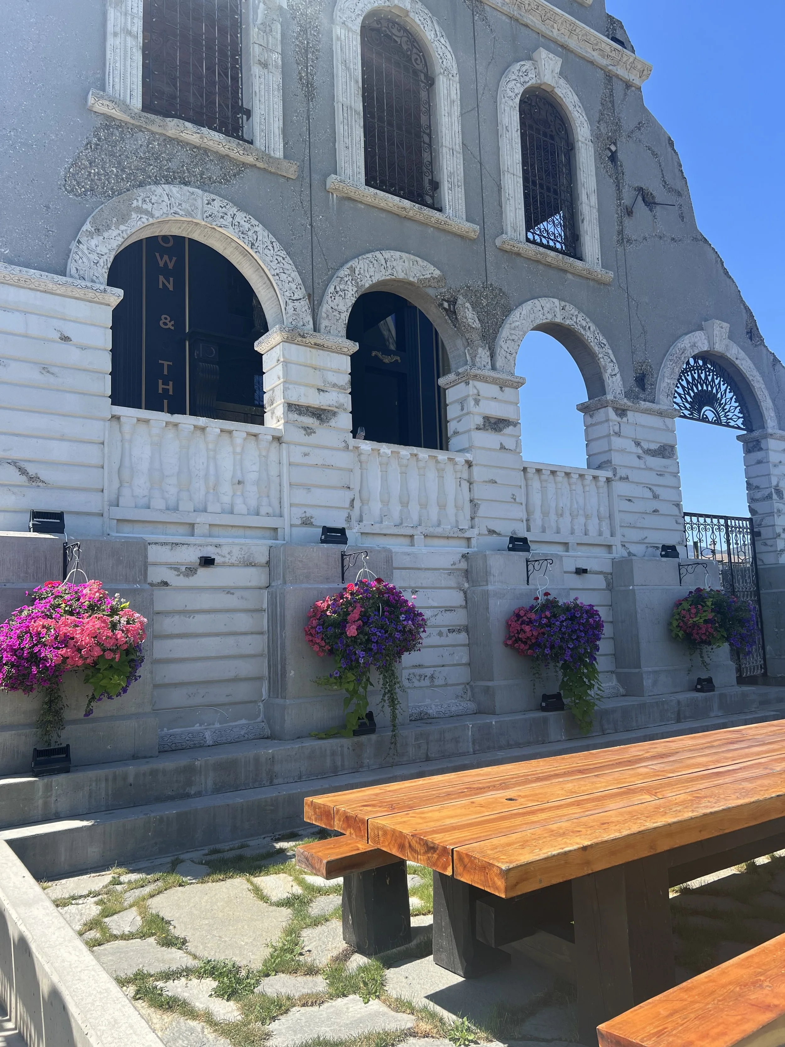 Old building with arched windows and ornate white stone detailing, decorated with hanging flower baskets, and a wooden picnic table in the foreground outside on a sunny day.