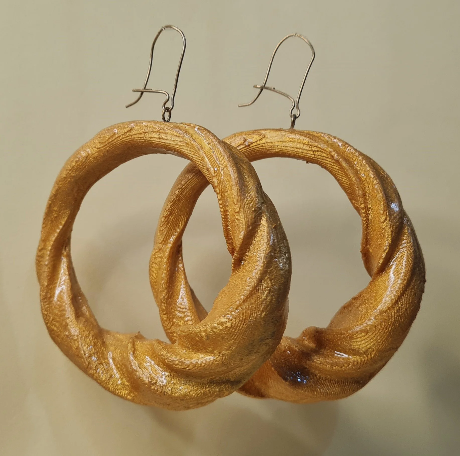 Un par de aretes dorados, impresos en 3D y luego pintados y con capa de resina. Diseño trenzado,  colgantes sobre un fondo neutro. Están diseñados con un estilo de aro grueso y tienen un gancho metálico para sujetarlos.