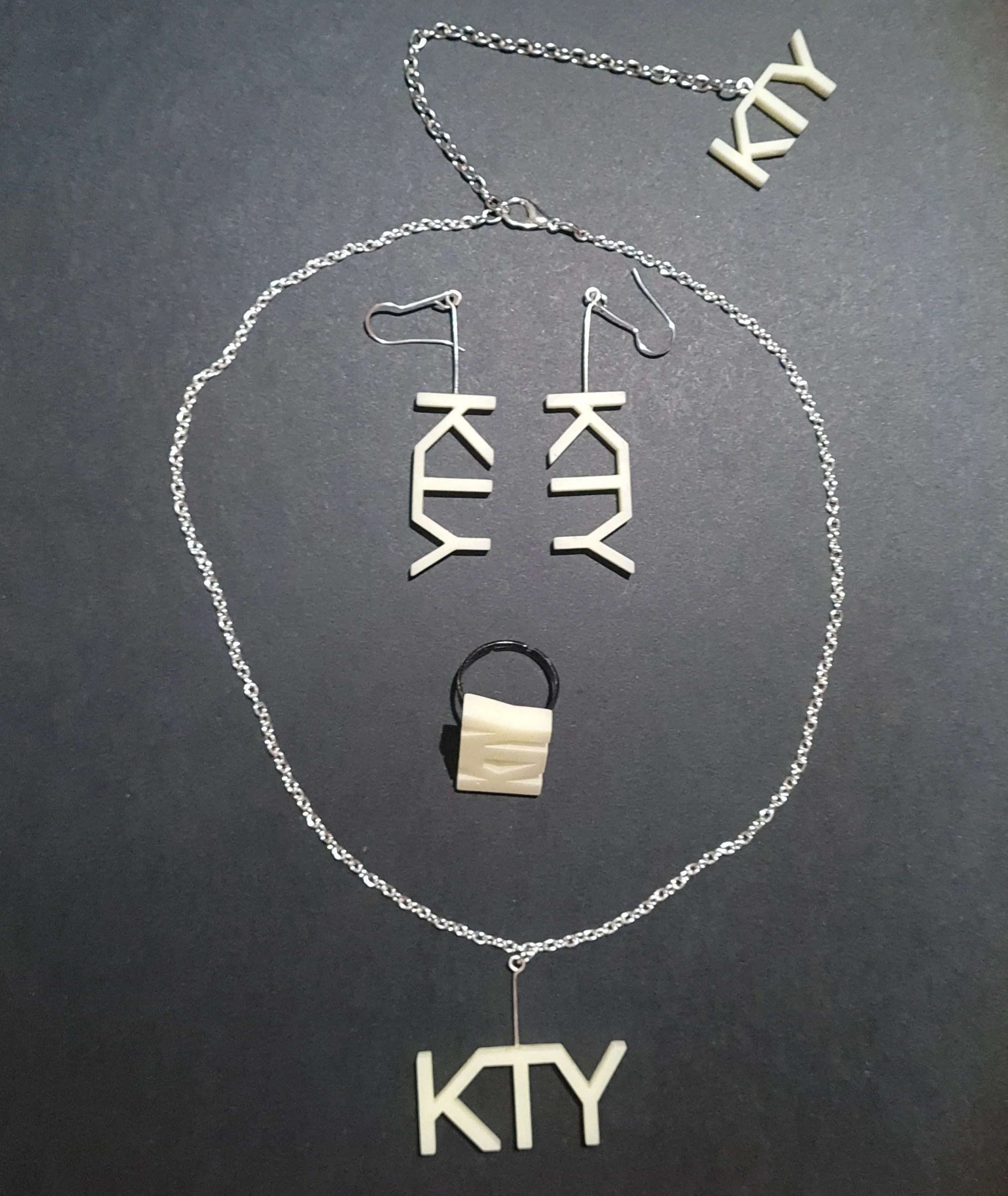 Conjunto de joyería con collares, pendientes y anillo, cada uno con las letras "KTY" en un diseño de resina blanca, montado en color plata, sobre fondo negro.