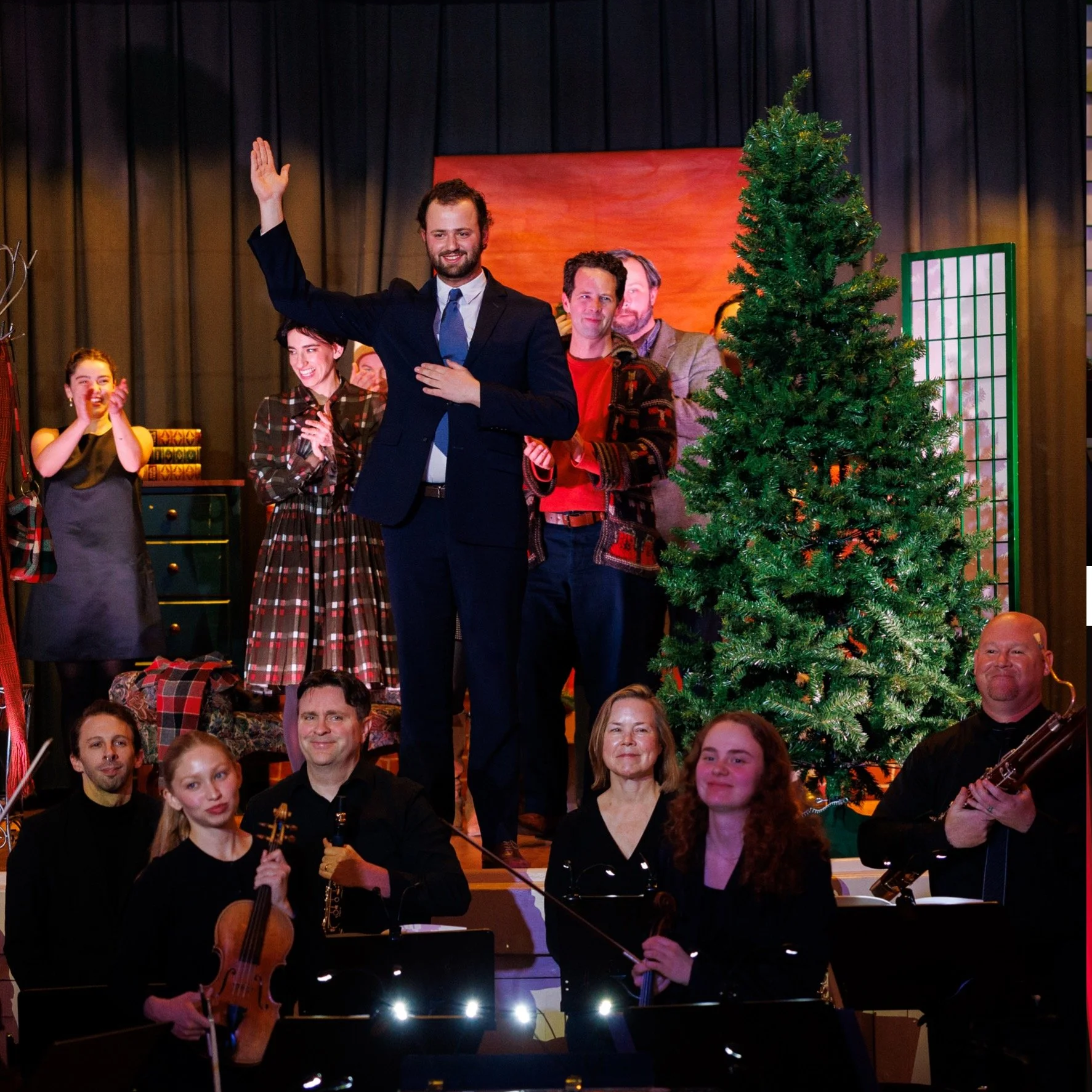 People on stage celebrating around a Christmas tree, some clapping and smiling, with musicians in front holding instruments.