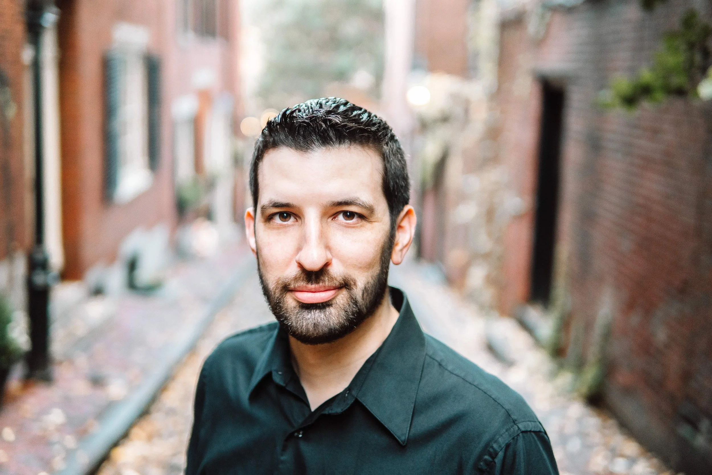 A man with dark hair and beard wearing a black shirt standing outdoors on a city street with brick buildings on either side, blurry background, fall leaves on the ground.