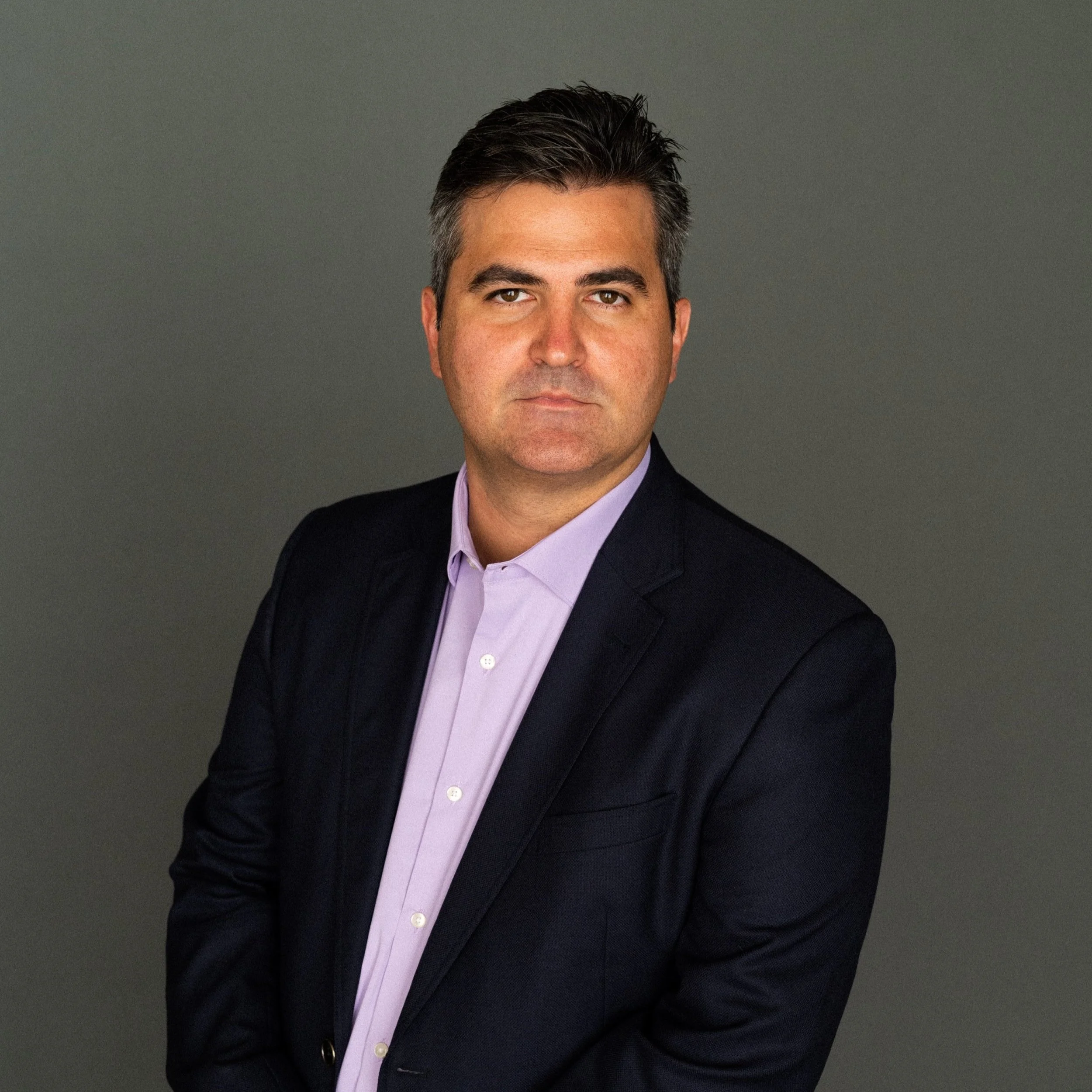 A man with short dark hair, wearing a dark suit jacket and a light purple shirt, posed against a plain gray background.