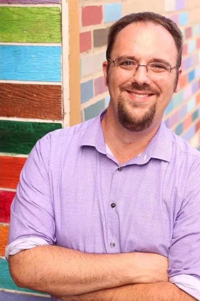 A smiling man with glasses and a beard, wearing a purple shirt, standing in front of a colorful wall made of painted wooden planks.