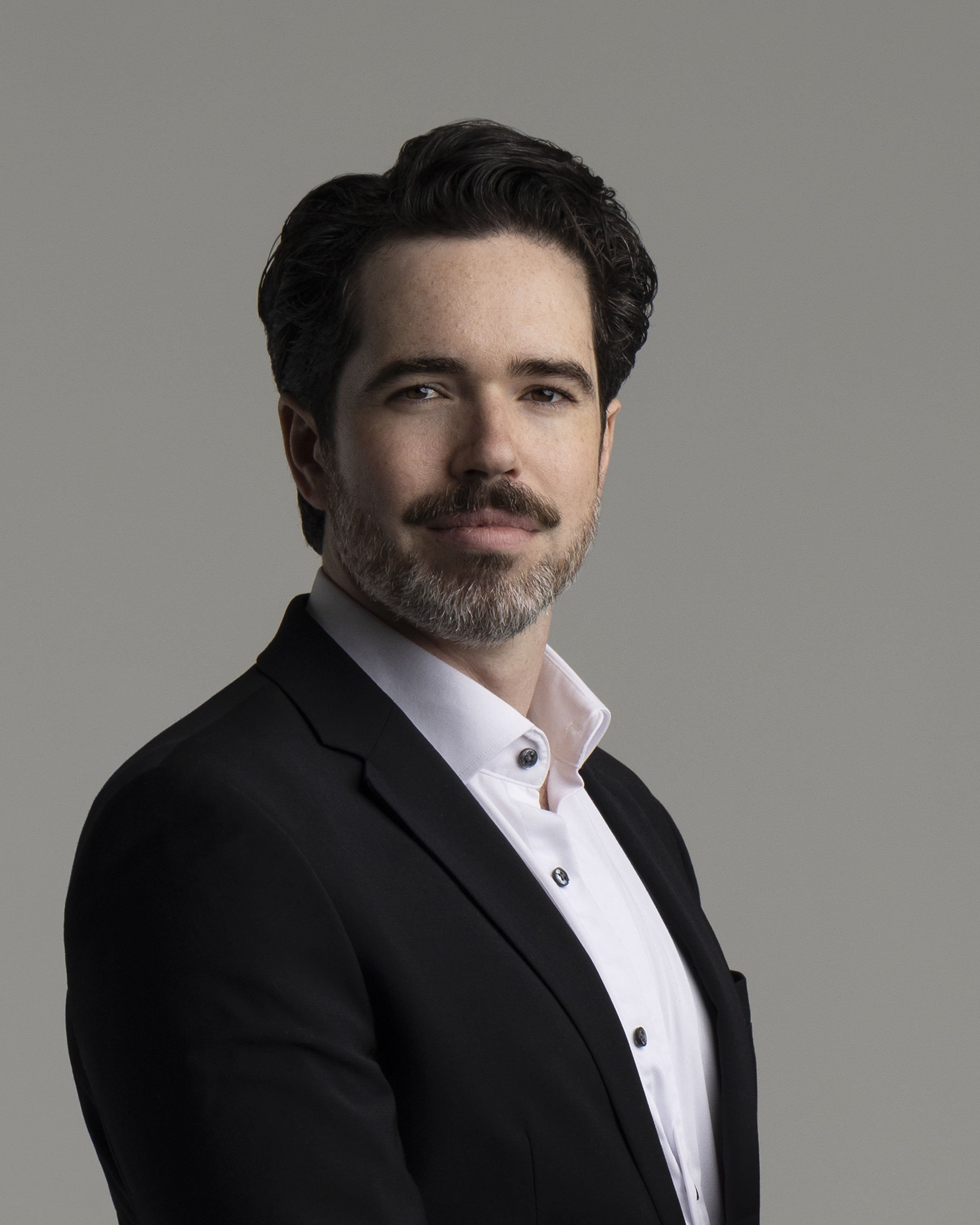 A man with dark hair and a beard wearing a black suit jacket and a white dress shirt looking at the camera with a neutral expression against a plain gray background.