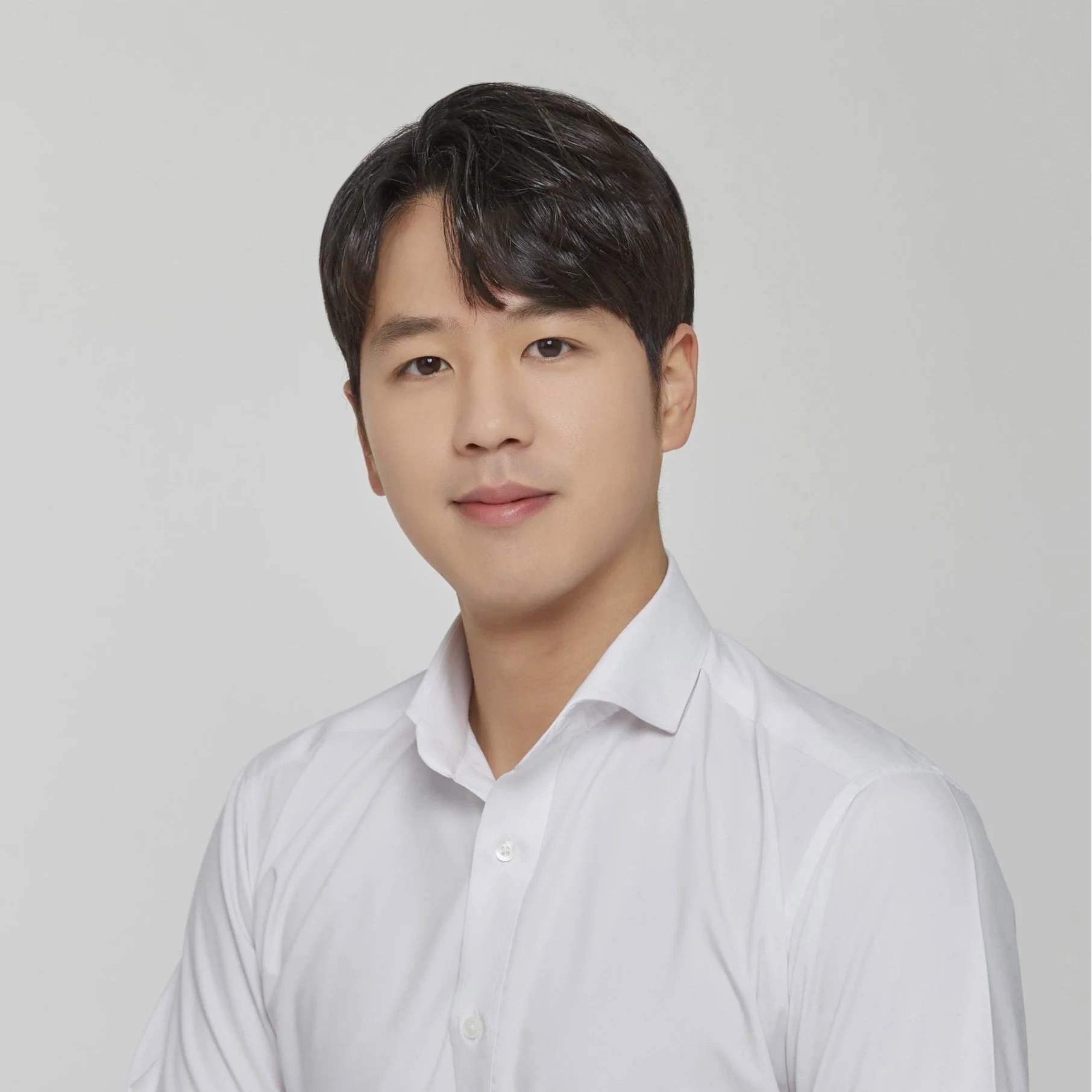 Portrait of a young man with dark hair, wearing a white dress shirt and looking at the camera with a subtle smile, against a plain light gray background.