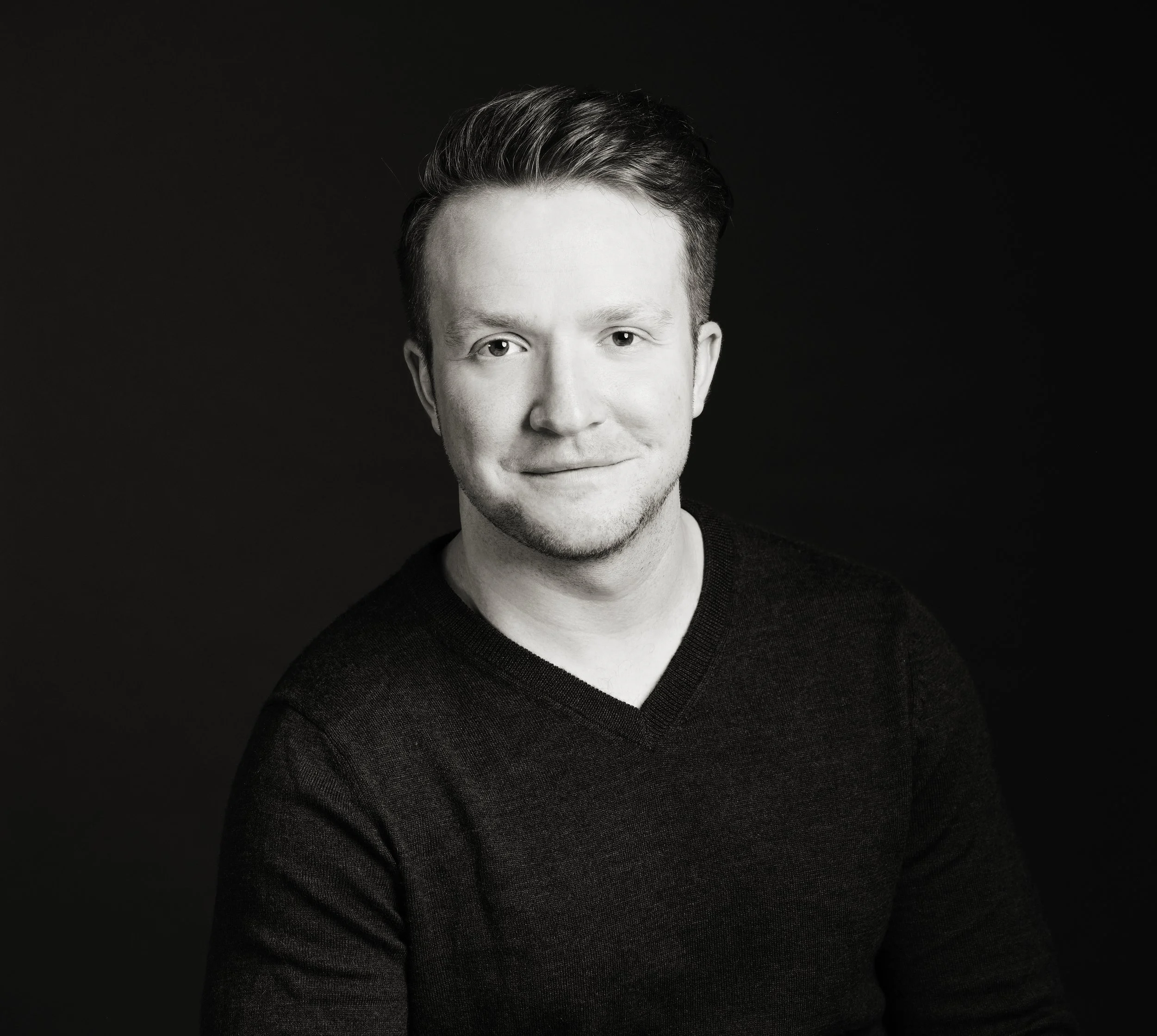 Black and white portrait of a young man with short, styled hair, smiling softly, wearing a dark V-neck sweater, against a solid dark background.