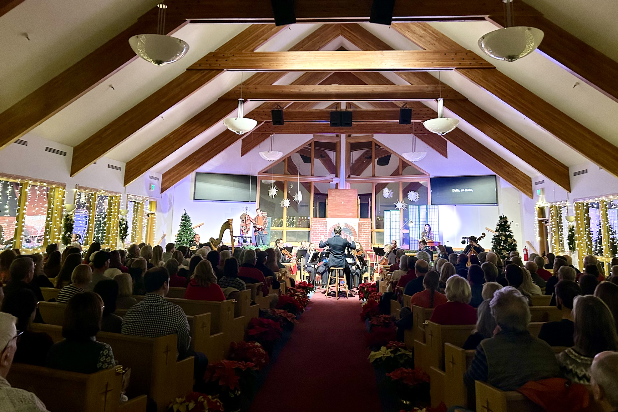 A Christmas concert in a church with a large audience, decorated with Christmas trees, poinsettias, and holiday lights, featuring a choir and an orchestra performing on stage under a peaked wooden ceiling.