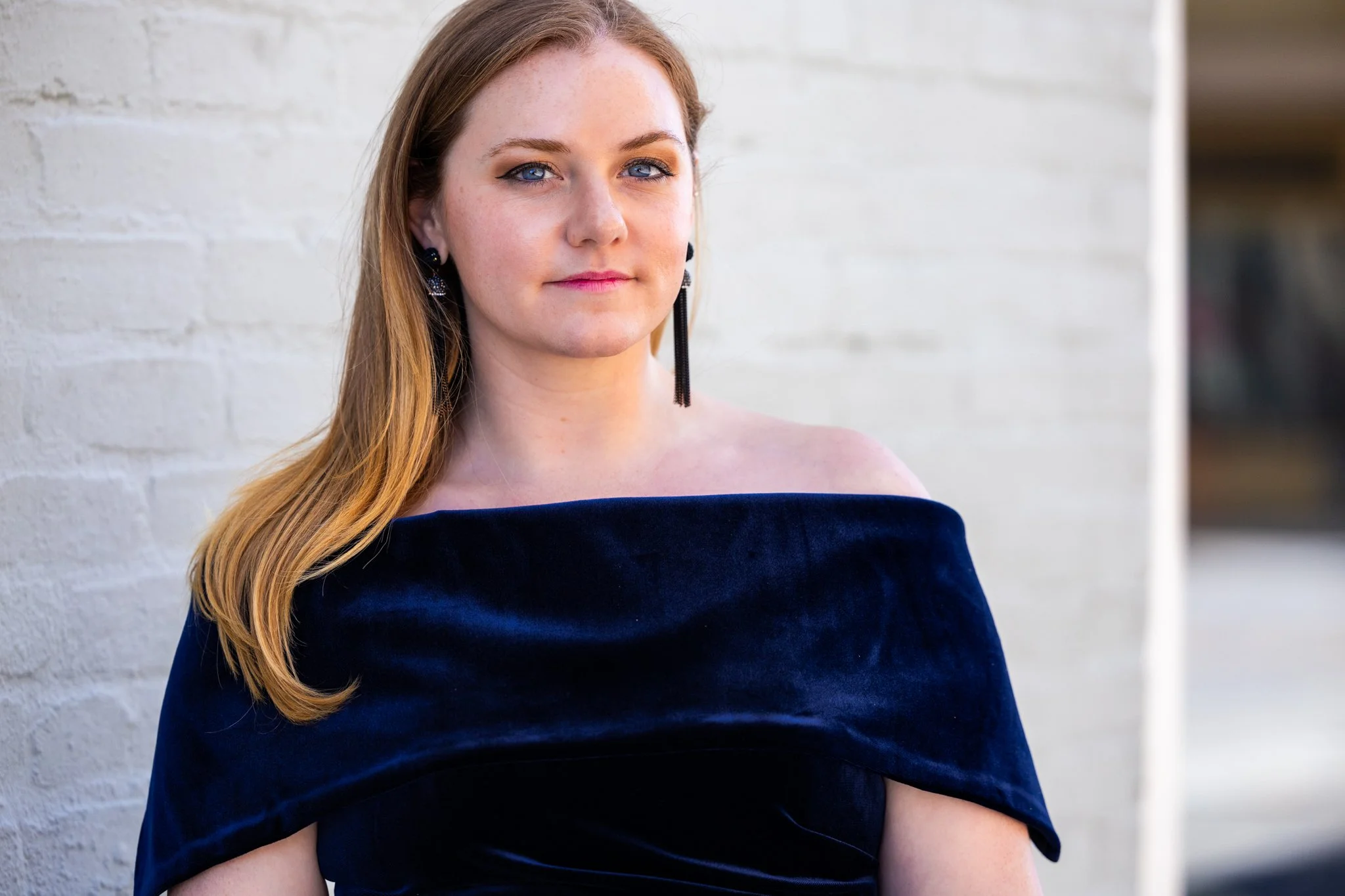 Portrait of a young woman with red hair, blue eyes, and fair skin, wearing a dark off-the-shoulder dress and black earrings, standing against a white brick wall.
