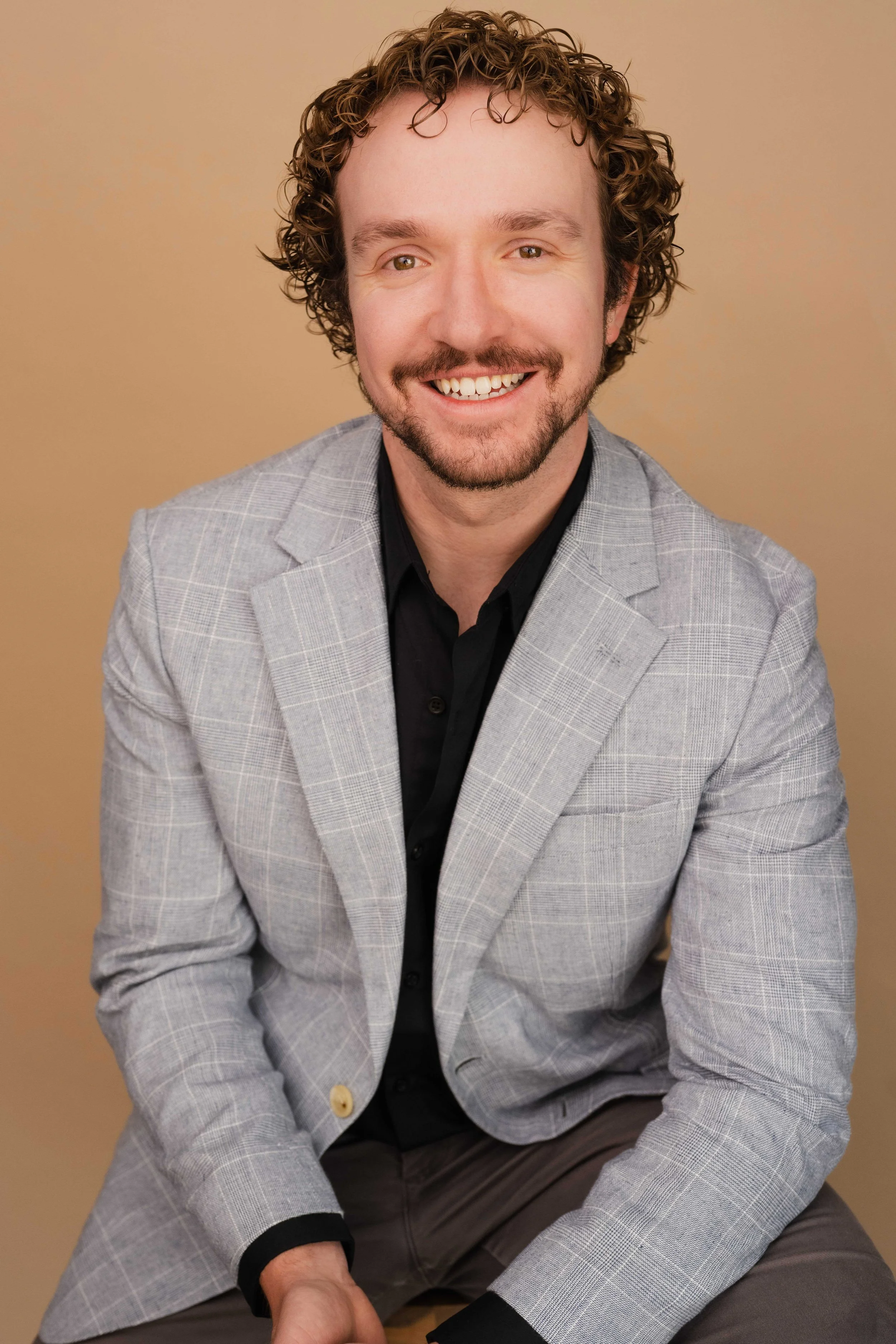 A man with curly hair, a beard, and a mustache, smiling in a gray checkered blazer and black shirt, sitting against a brown background.