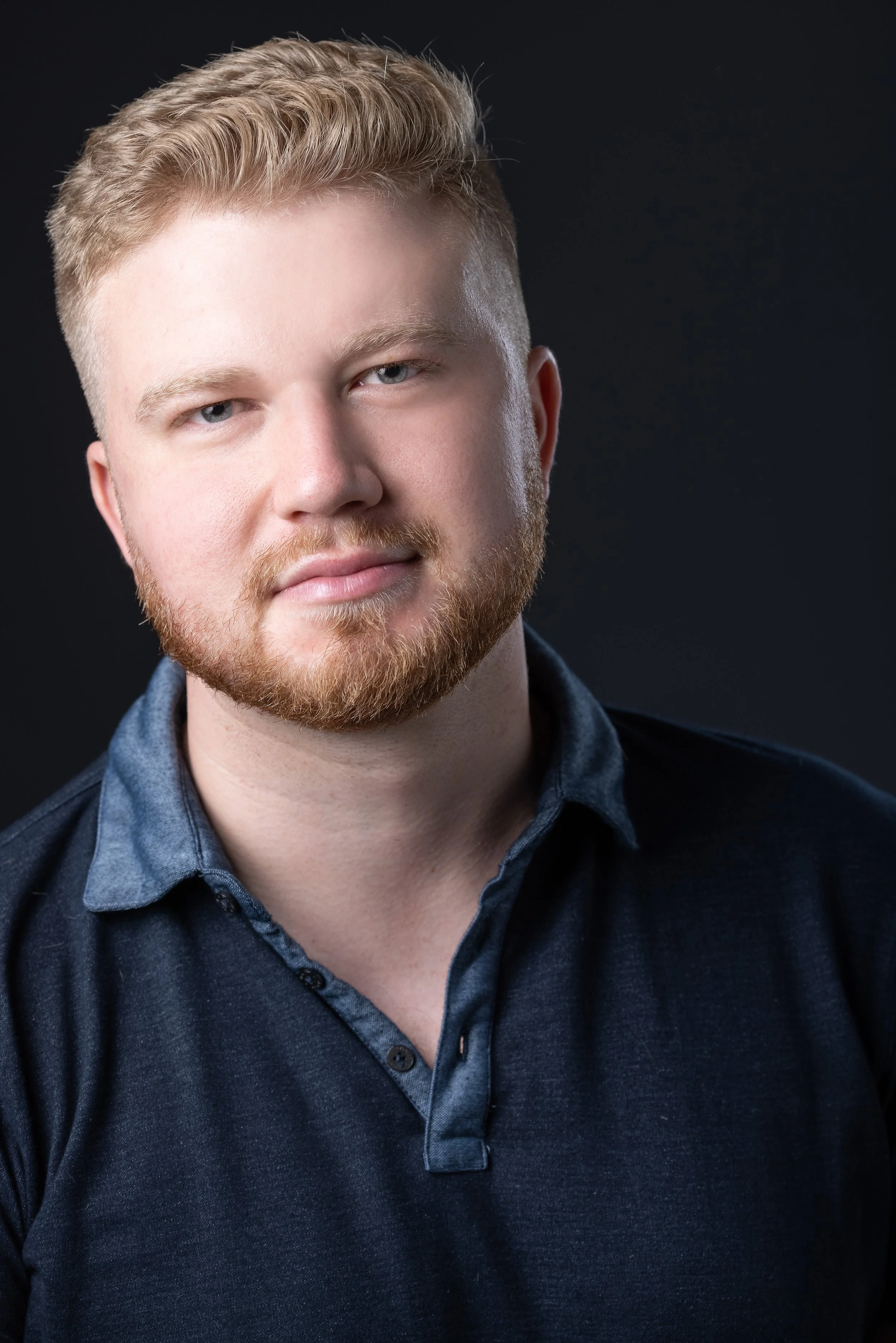 A portrait of a young man with light skin, blonde hair, a beard, and blue eyes wearing a dark-colored collared shirt against a black background.
