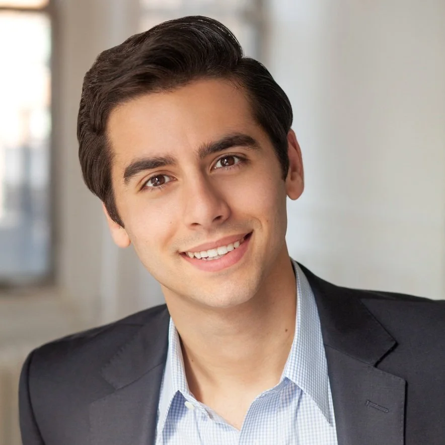 Headshot of a young man in a business suit, smiling, with a blurred office background.