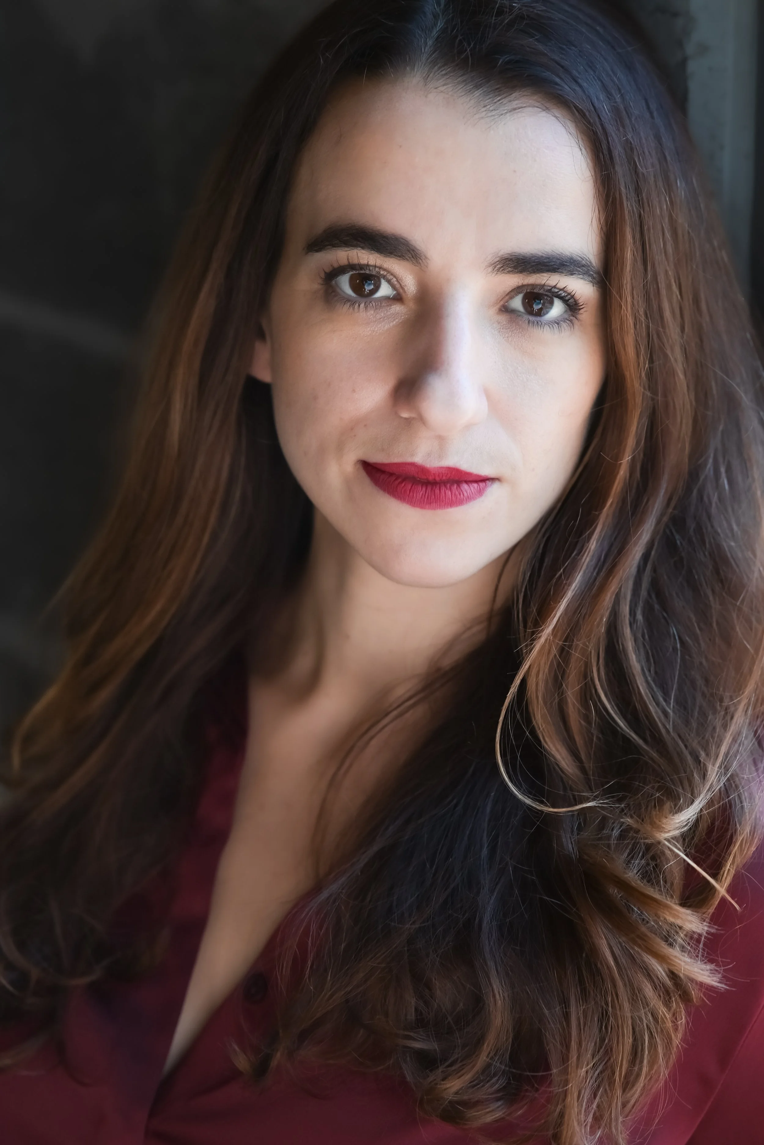A close-up portrait of a woman with long, wavy brown hair, brown eyes, red lipstick, and a deep burgundy blouse.
