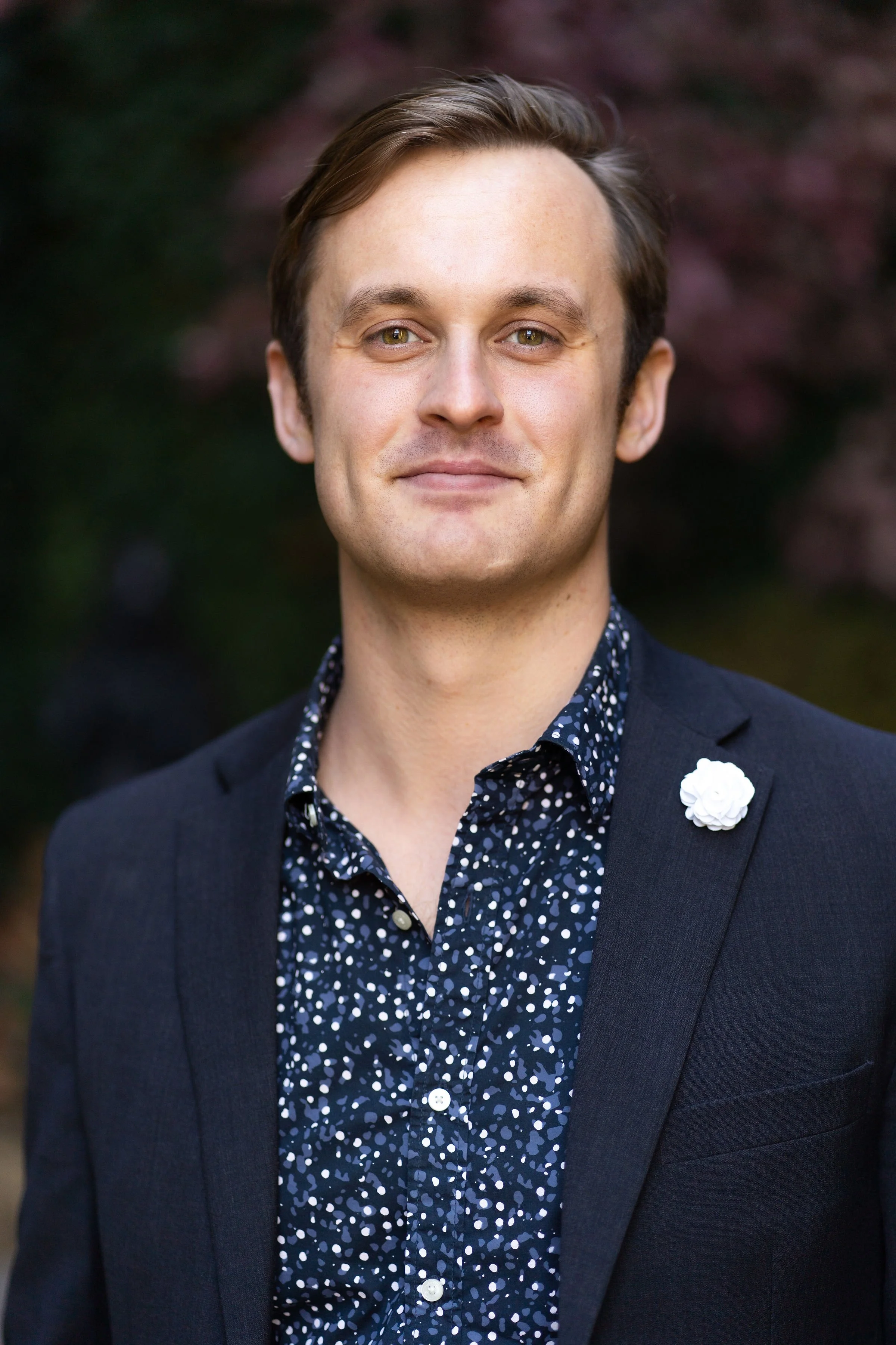 A man with light skin and brown hair smiling outdoors, wearing a dark blazer with a white flower pin and a navy shirt with white and blue dots, with blurred trees in the background.