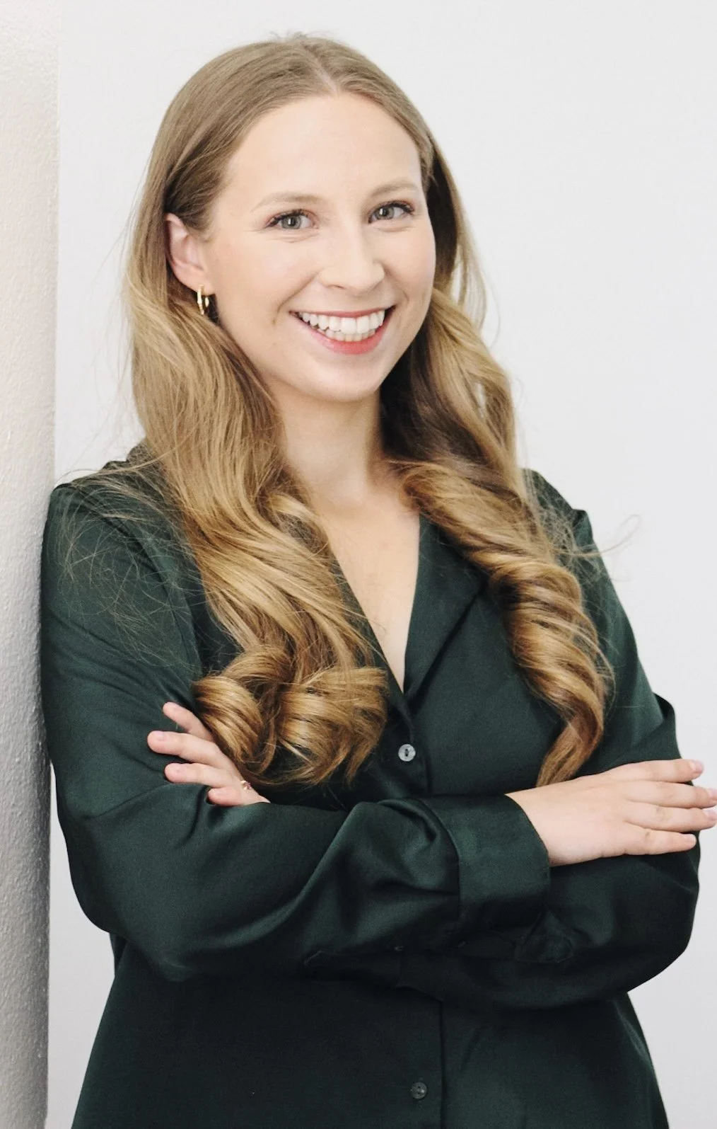 A woman with long, wavy red hair, wearing a dark green satin shirt, smiling with arms crossed, standing against a light-colored wall.