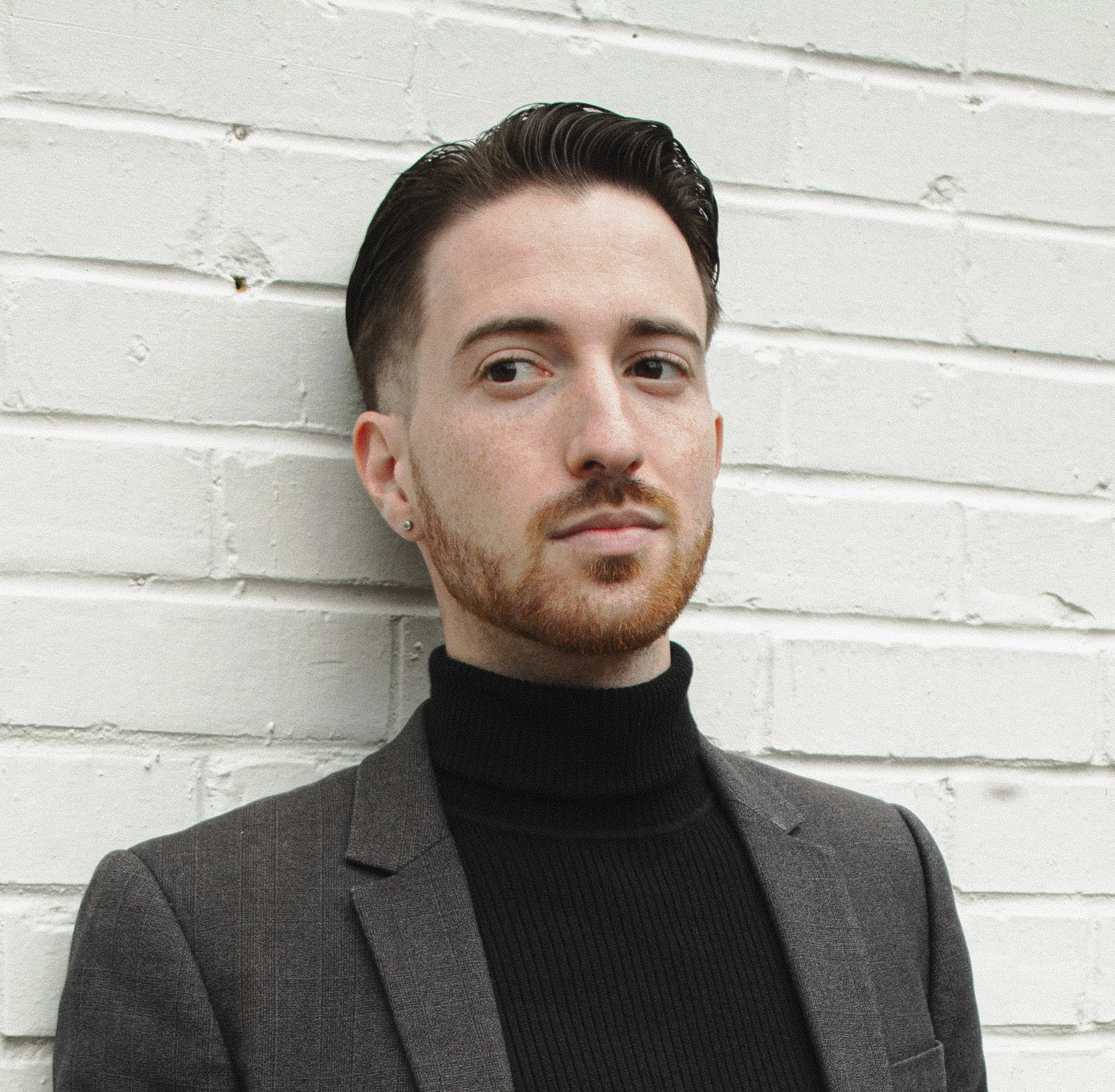 A man with a black turtleneck and grey blazer standing against a white brick wall.