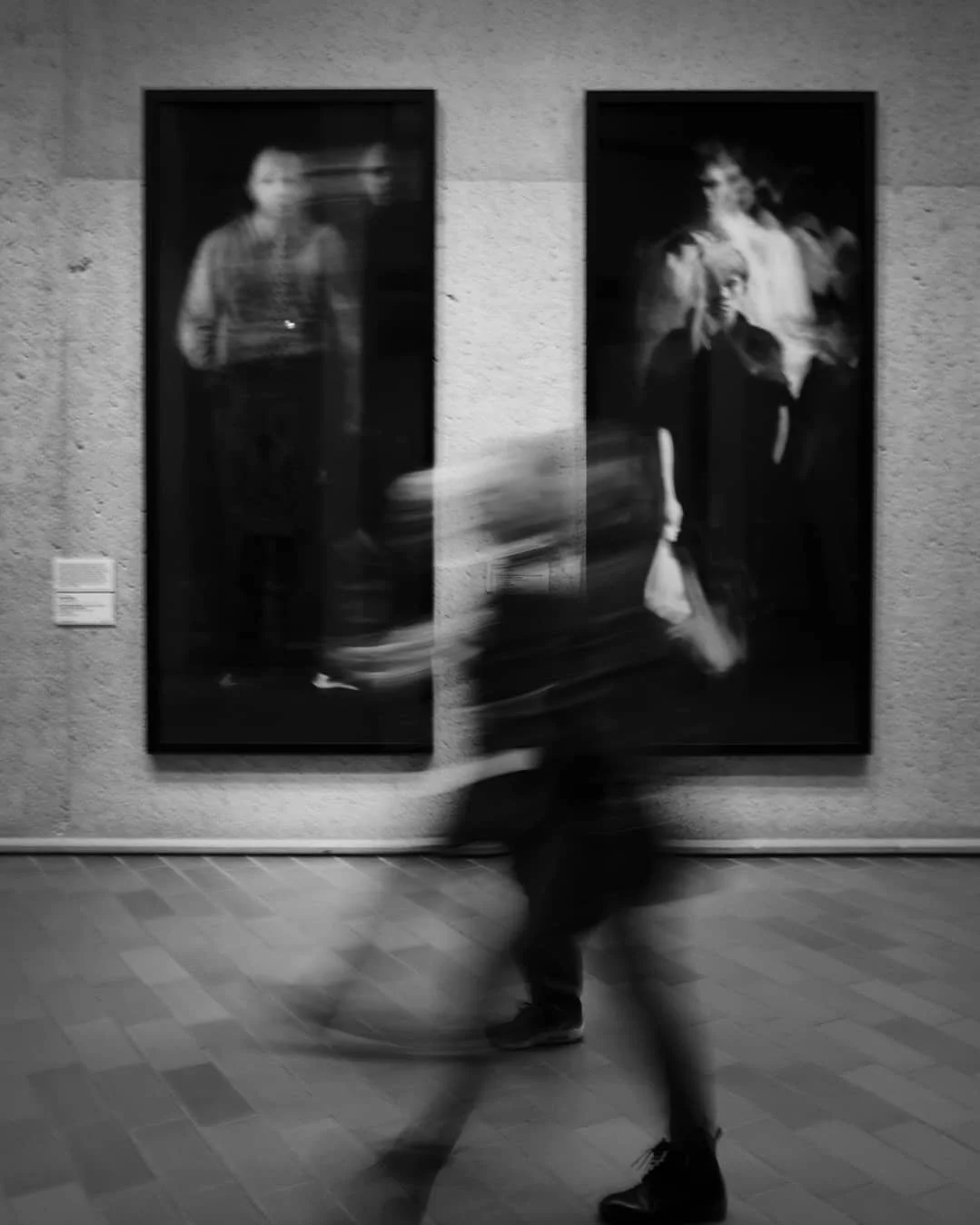Blurred motion of a person walking past two abstract art pieces on a wall.