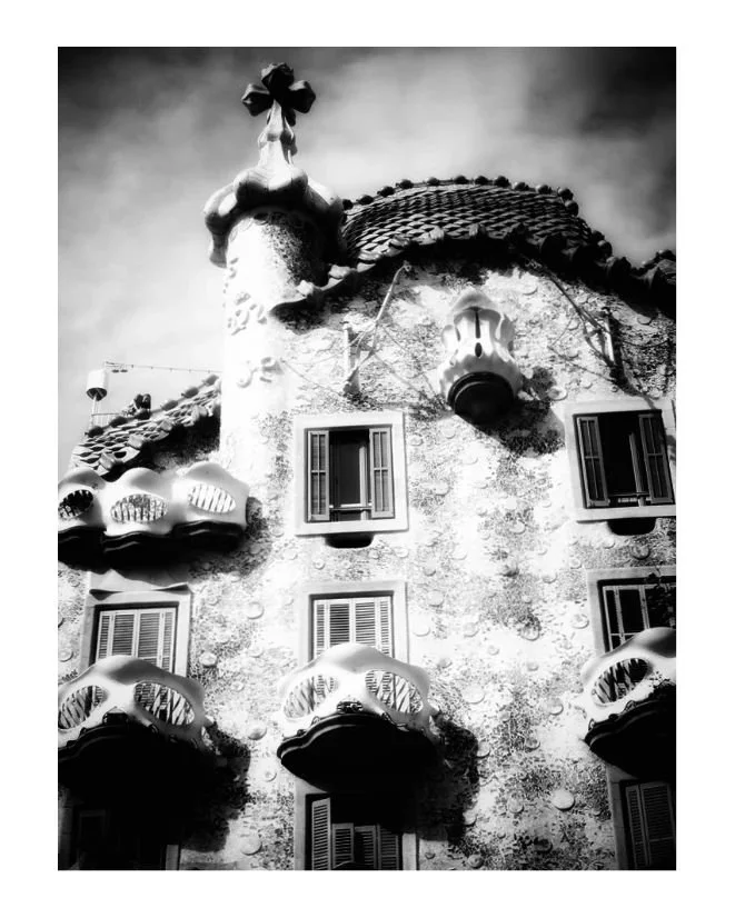 Black and white image of an ornate building with unique wavy balconies and a textured facade, featuring an undulating roof and a cross-shaped structure on top.
