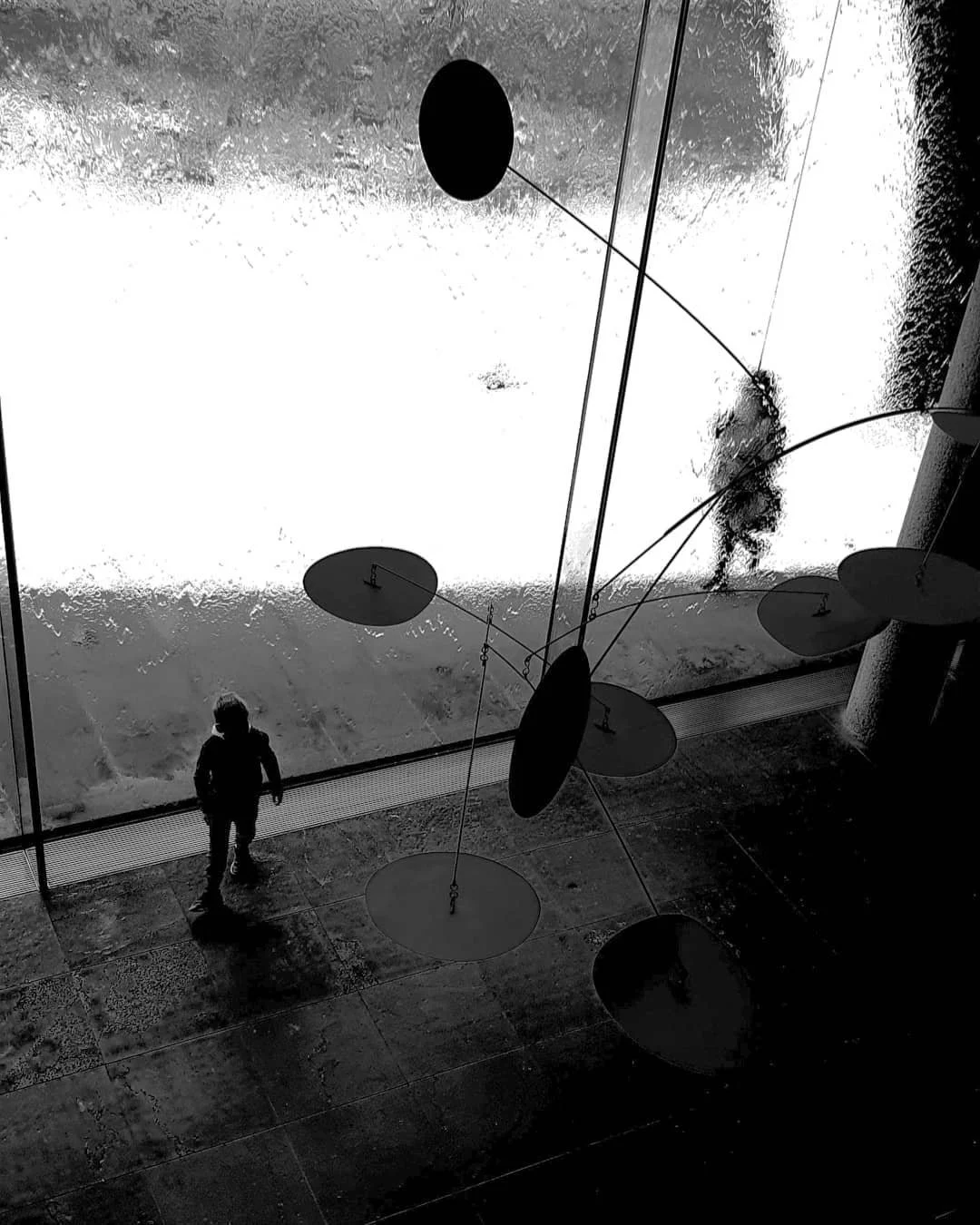 Black and white image of a silhouette of a person walking beside large abstract mobile sculptures with circular elements, next to a bright window.