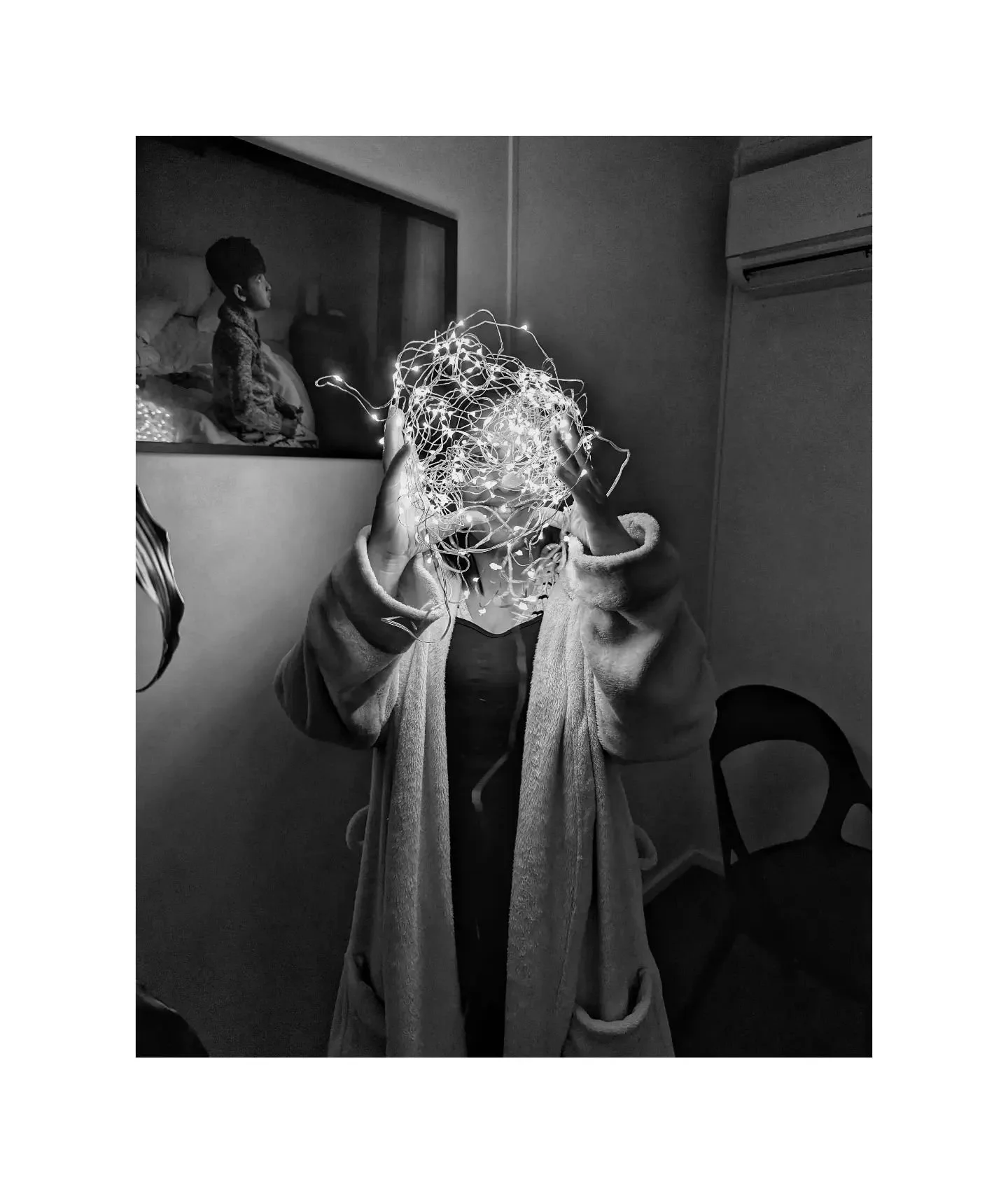 Person holding string lights in front of face, wearing a robe, with a chair and wall art in the background.