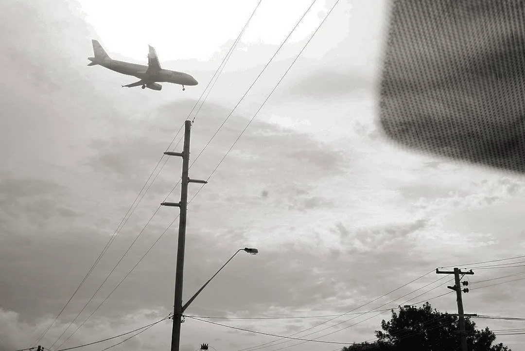 Airplane flying over utility poles against a cloudy sky, with power lines visible.