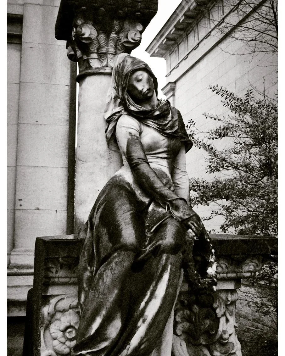 Black and white photo of a stone statue of a seated woman draped in cloth, leaning against a decorative column, with architectural details and foliage in the background.