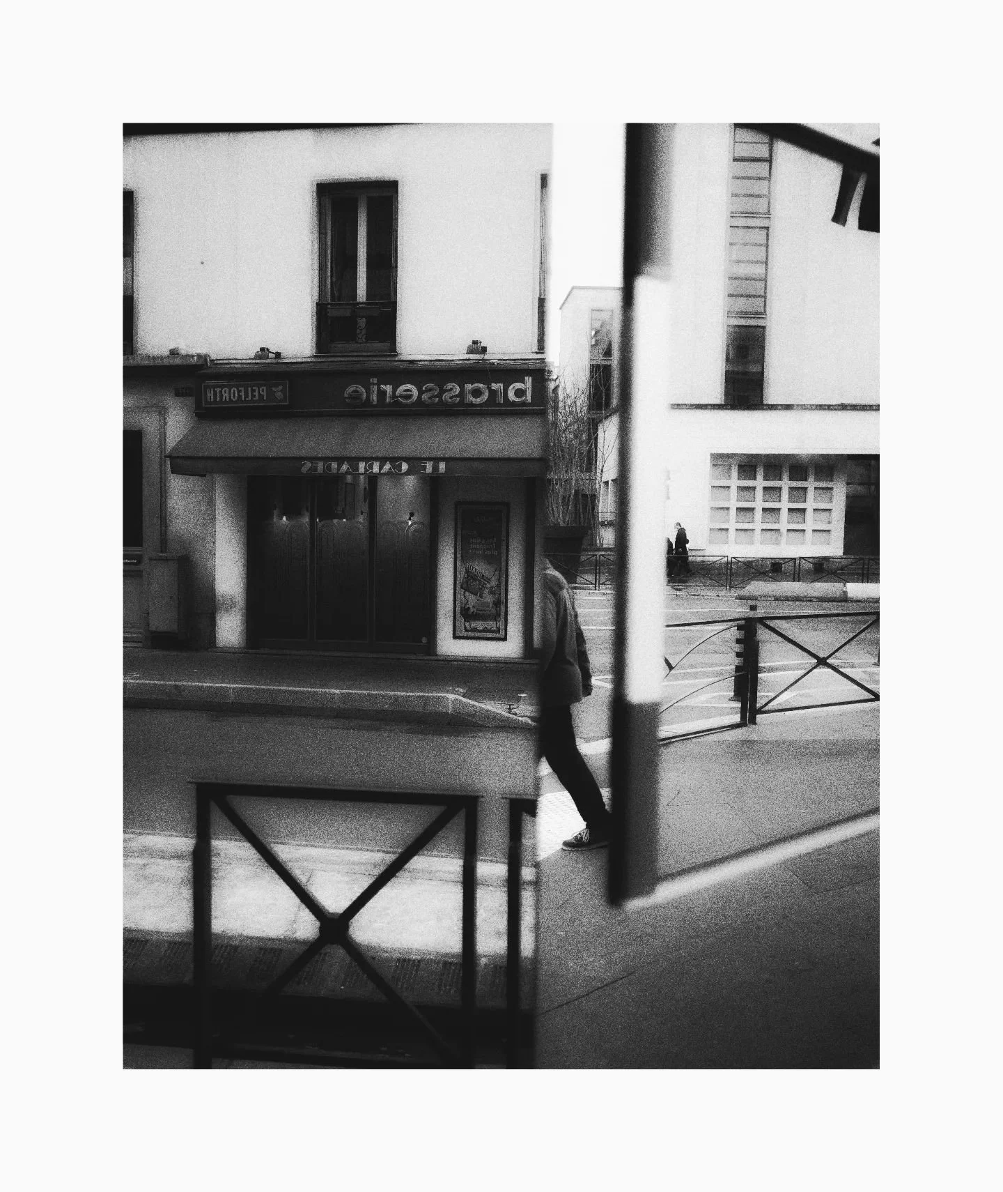 Black and white photograph of a street scene with a solitary person walking on the sidewalk in front of a building with a large awning. The building has signage that is partially visible and appears to be a cafe or similar establishment. The scene is